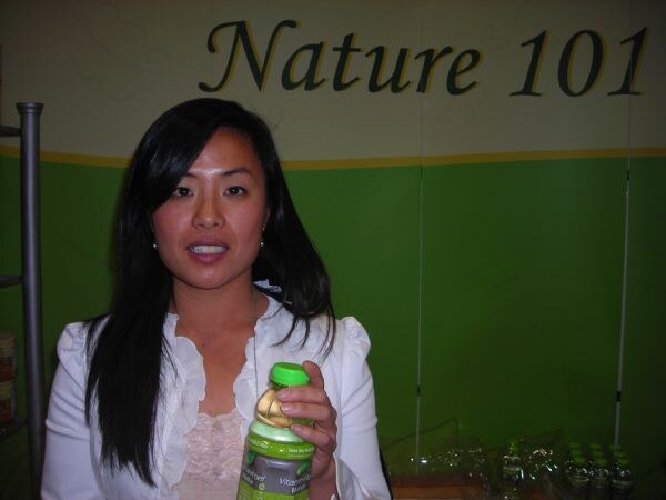A woman with long dark hair holds a green-capped bottle of vitamin and fiber water in front of a green and cream Nature 101 sign.