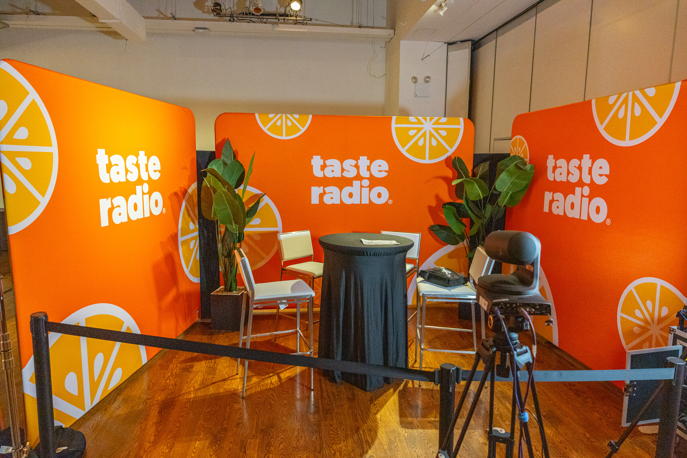 Bright orange Taste Radio backdrop with two chairs, a round table, plants and camera equipment behind a rope line.