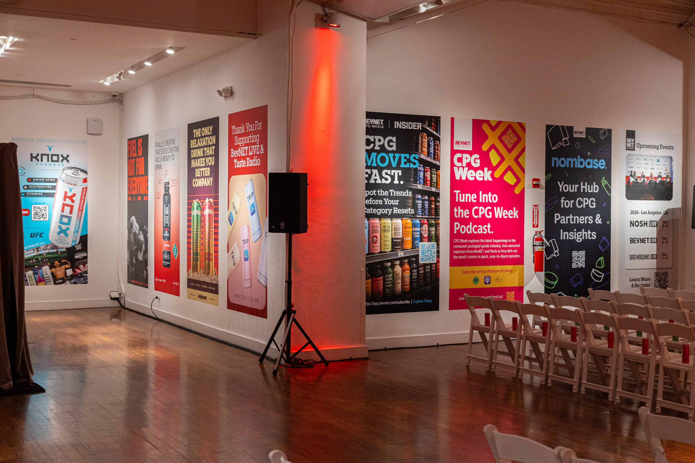 Wall of posters for CPG Week, Nombase and upcoming events beside a speaker stand and rows of chairs.