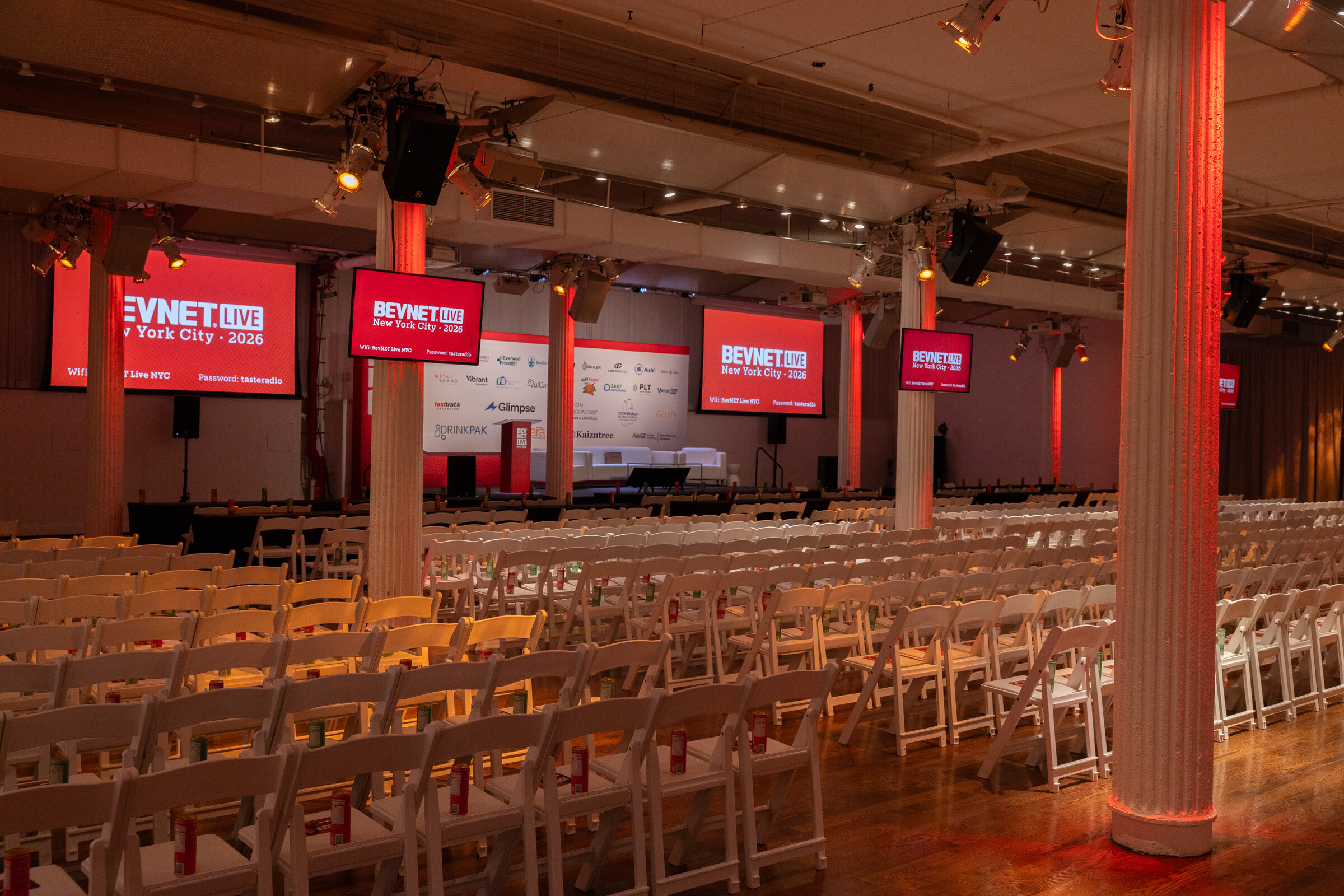Wide view of the main hall with red-lit columns, BevNET Live screens and rows of white chairs.