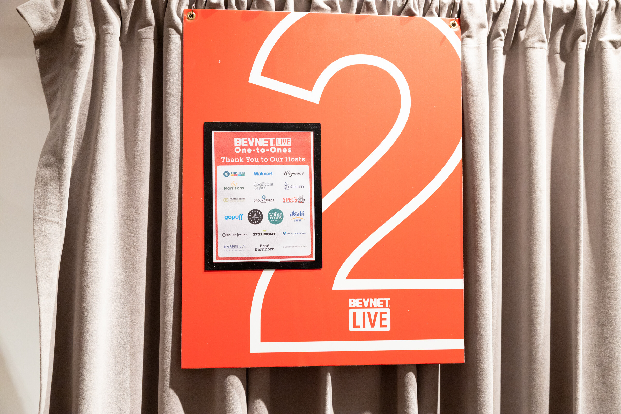 Red booth sign with a large number 2 and a list of One-to-One hosts pinned to a gray curtain.