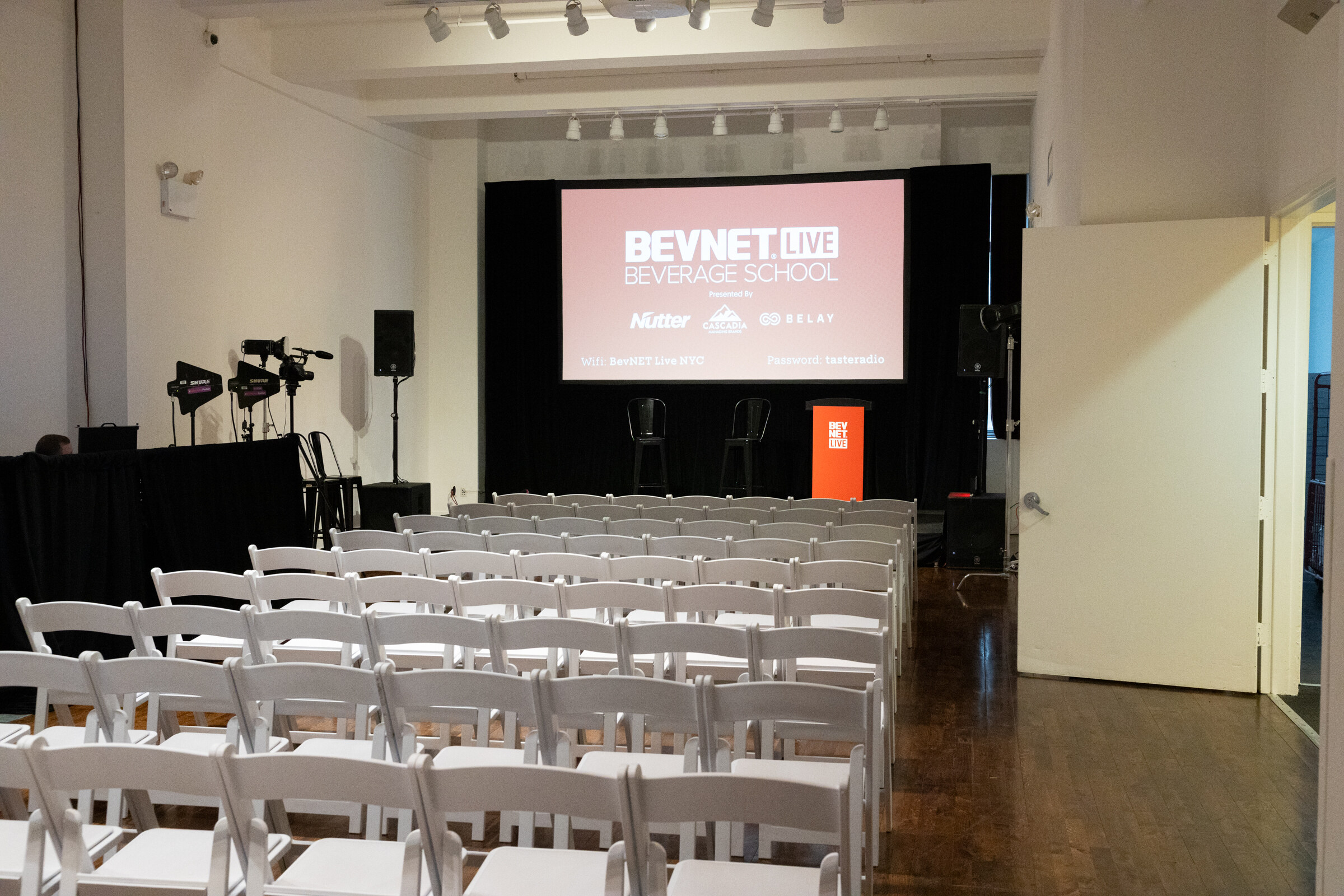 Small room with rows of white chairs facing a screen showing BevNET Live Beverage School and a red podium.