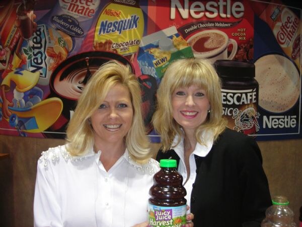 Two smiling blonde women pose in front of a Nestle mural showing Nesquik, Coffee-mate, Nescafé and Nestea; one holds a bottle of Juicy Juice.