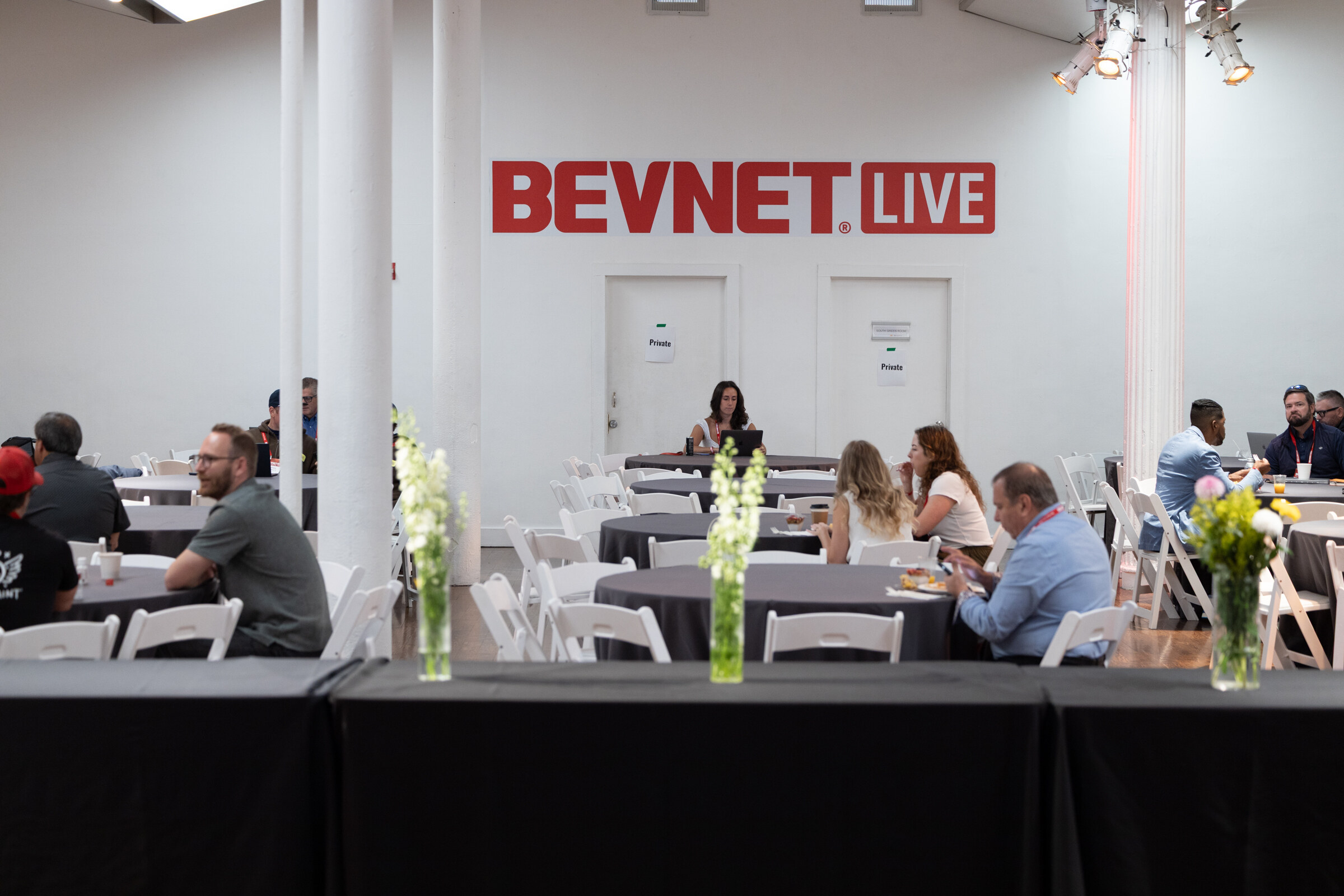 Attendees at round gray tables with flower centerpieces in a white room with a red BevNET Live wall sign.