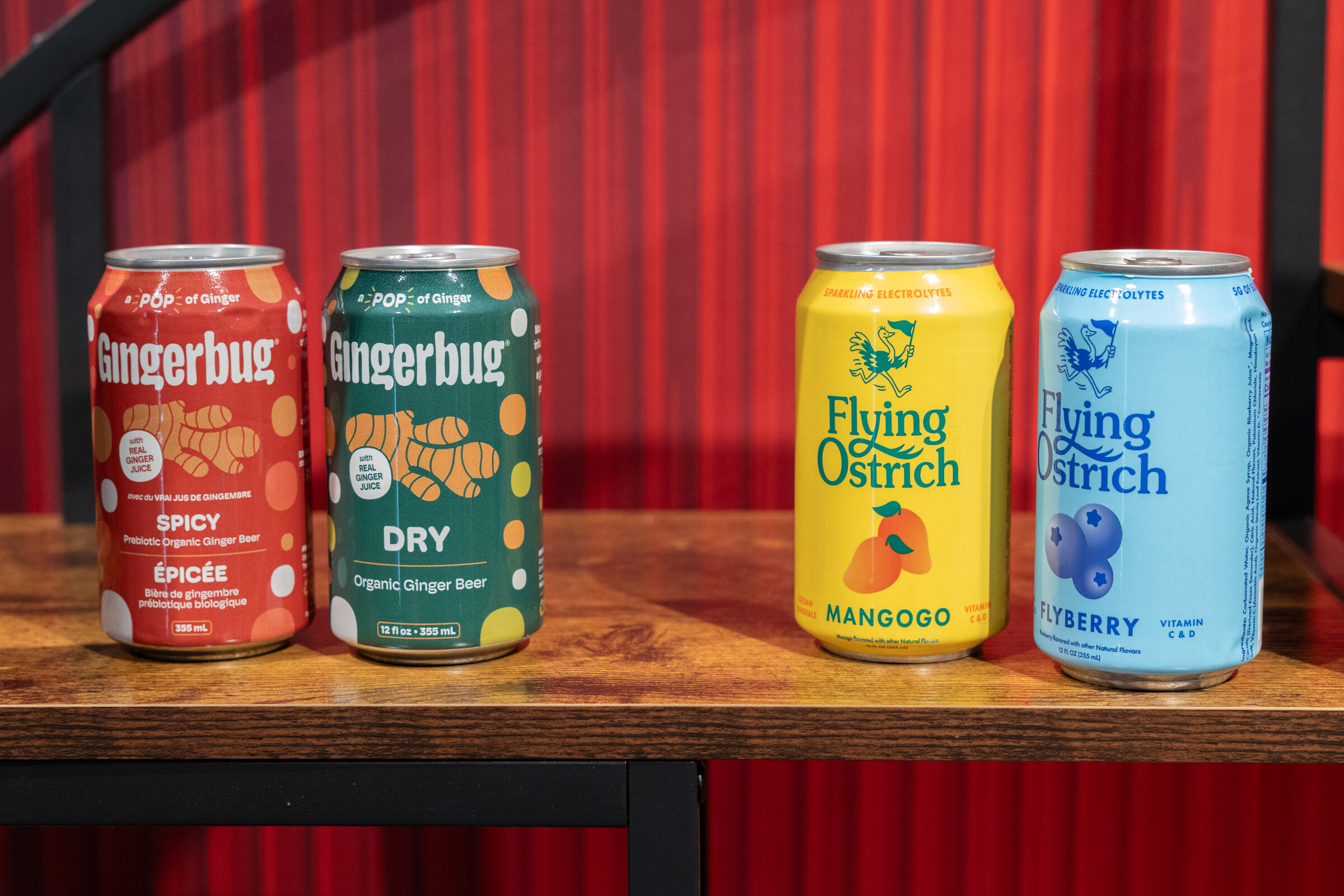 Cans of Gingerbug Spicy and Dry and Flying Ostrich Mangogo and Blueberry on a wooden shelf against red.
