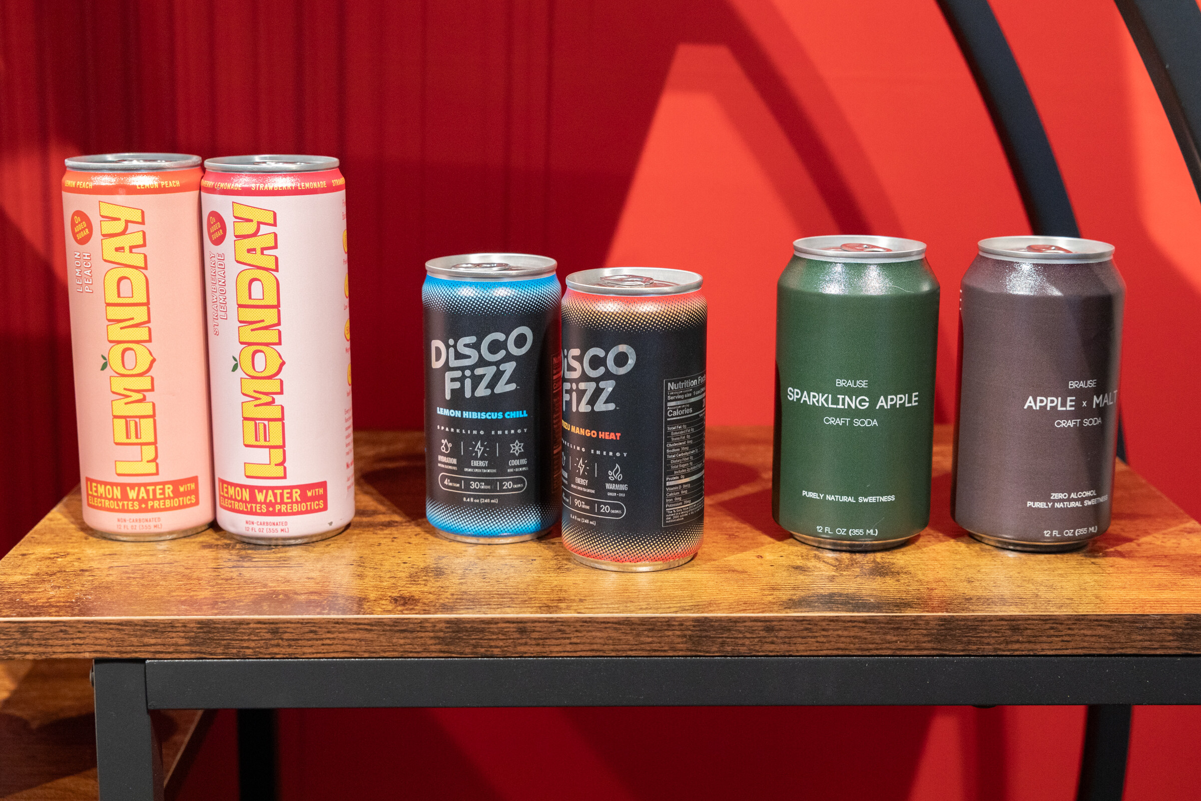 Cans of Lemonday, Disco Fizz and Brause craft soda lined up on a wooden shelf against red.