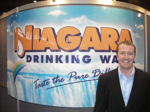 A smiling man in a dark blazer and light blue shirt stands in front of a large Niagara Drinking Water sign showing a waterfall and rainbow.