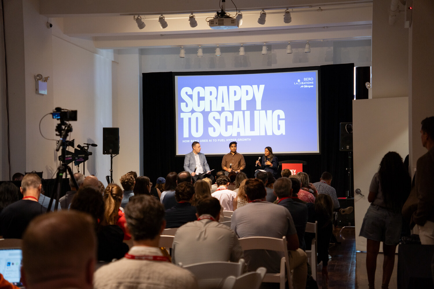Wide view of a seated audience facing a stage where three panelists sit beneath a purple “Scrappy to Scaling” slide.