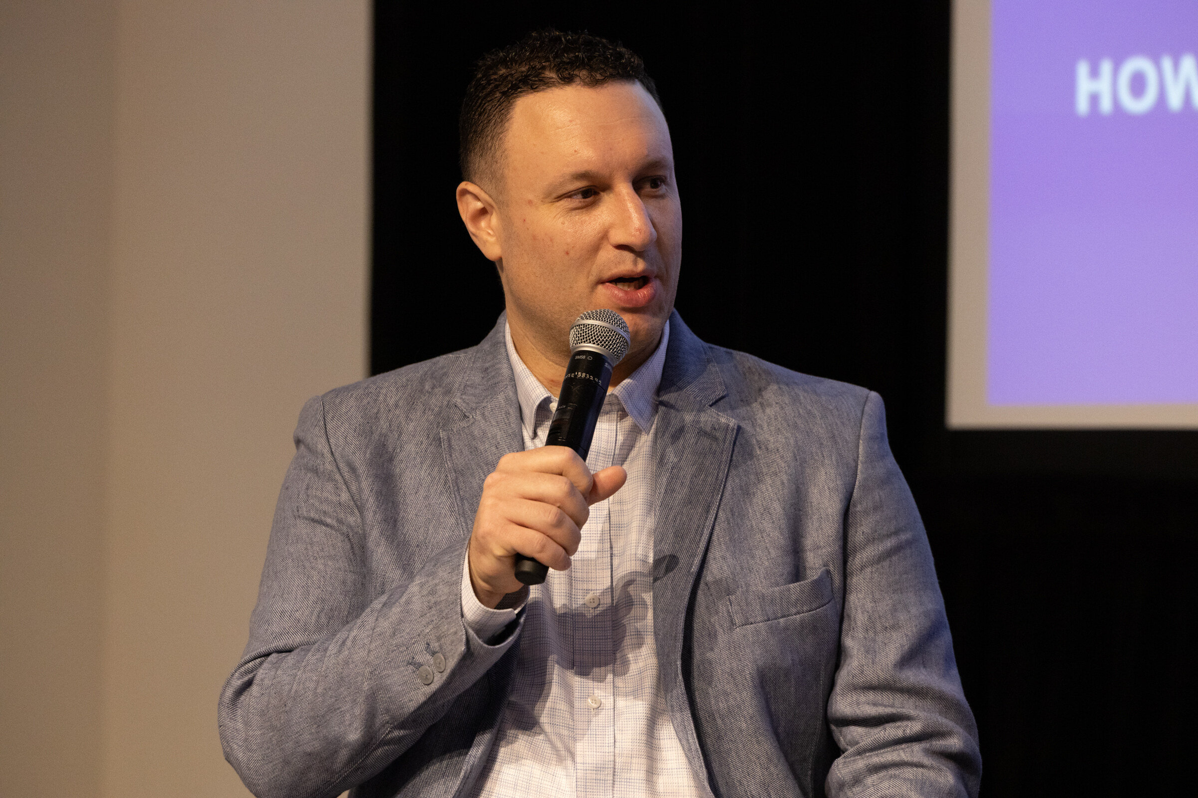 Man in a grey blazer speaking into a microphone on stage, a purple slide behind him.