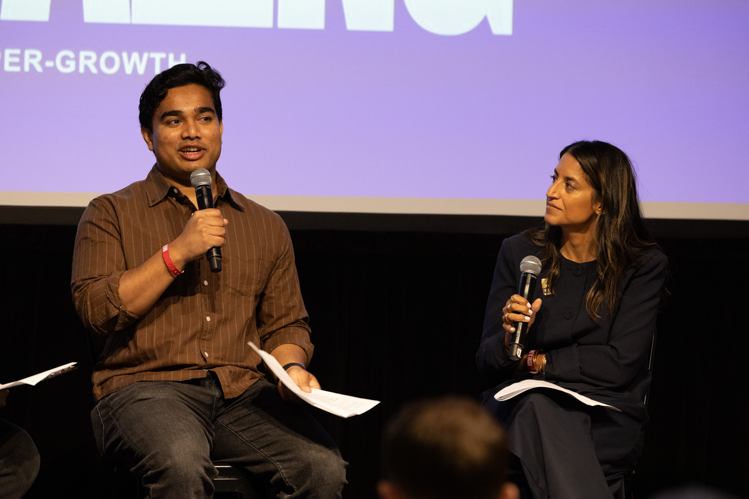 Man in a brown striped shirt speaking into a microphone while a woman beside him listens, a purple slide behind them.