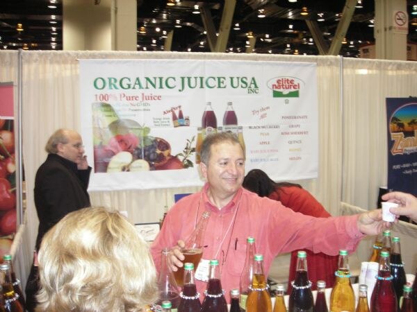 A man in a pink shirt reaches out with a sample cup at the Organic Juice USA booth, with Elite Naturel juice bottles on the counter and a banner listing fruit flavors behind him.