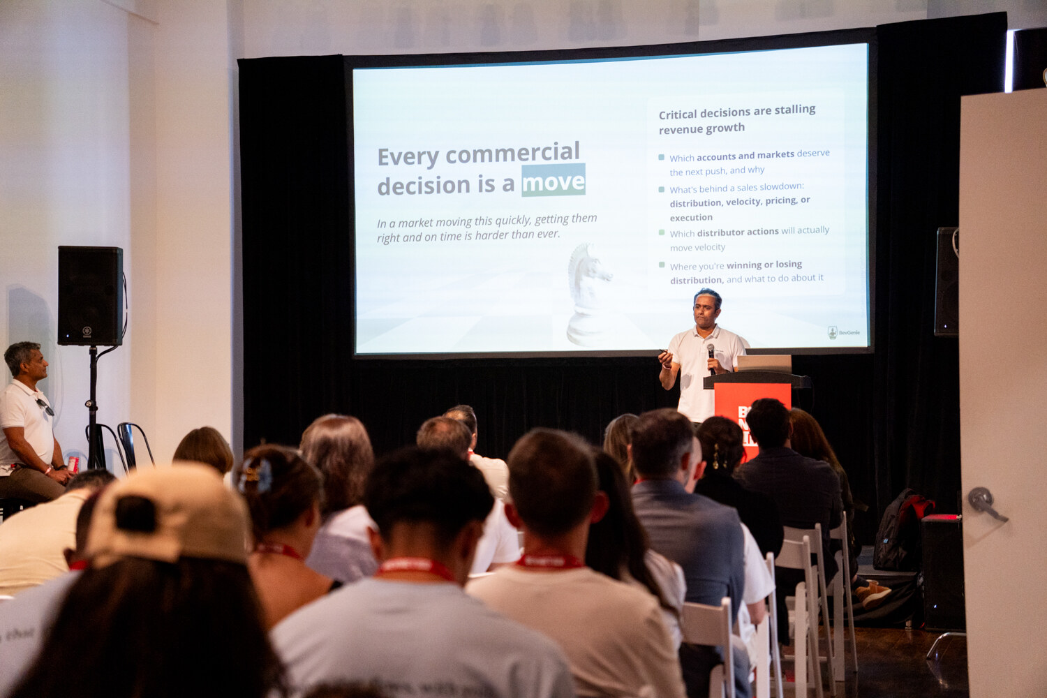 Speaker at a podium before a slide reading “Every commercial decision is a move,” seen over a seated audience.