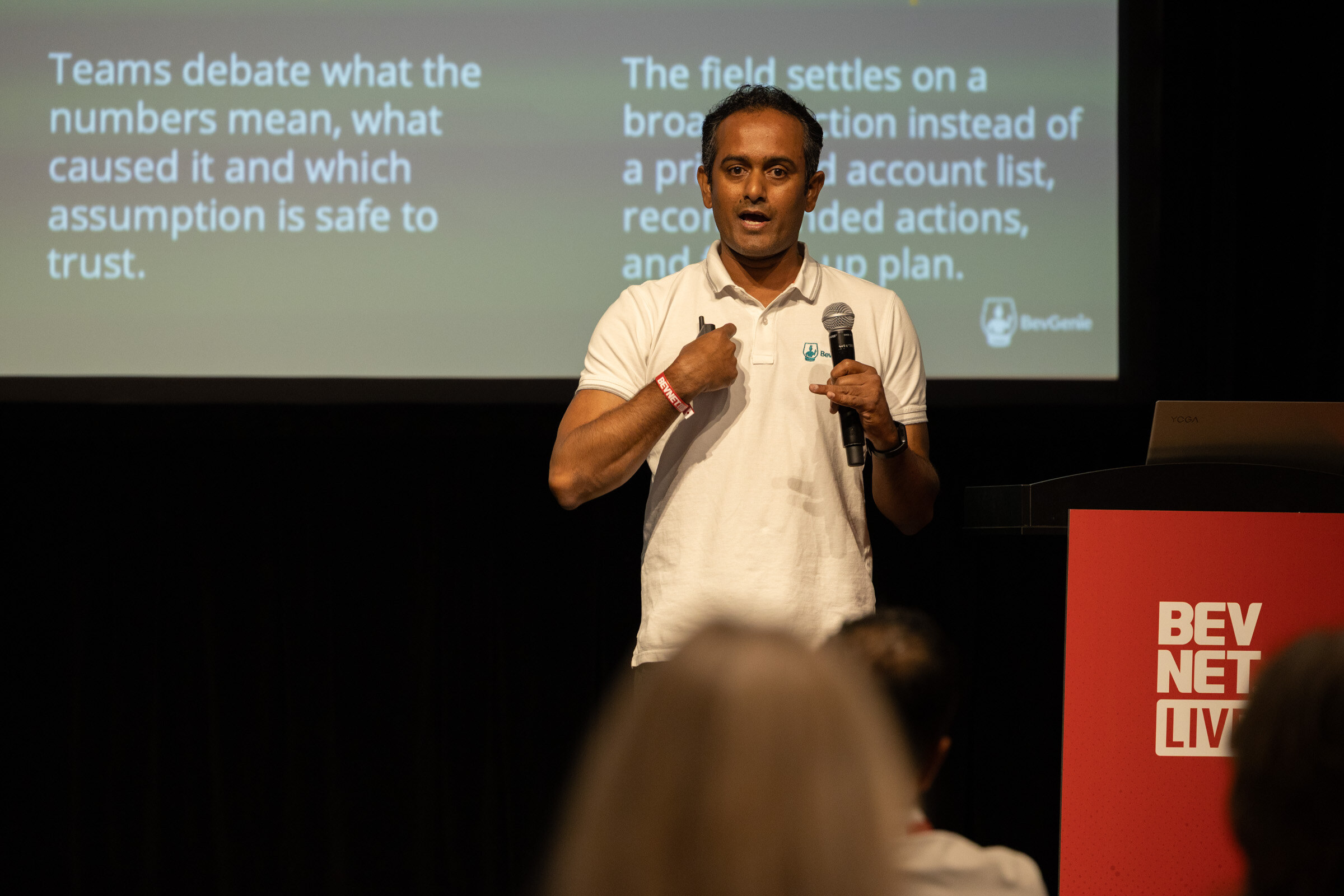 Man in a white polo speaking into a microphone in front of a slide about teams debating what the numbers mean.