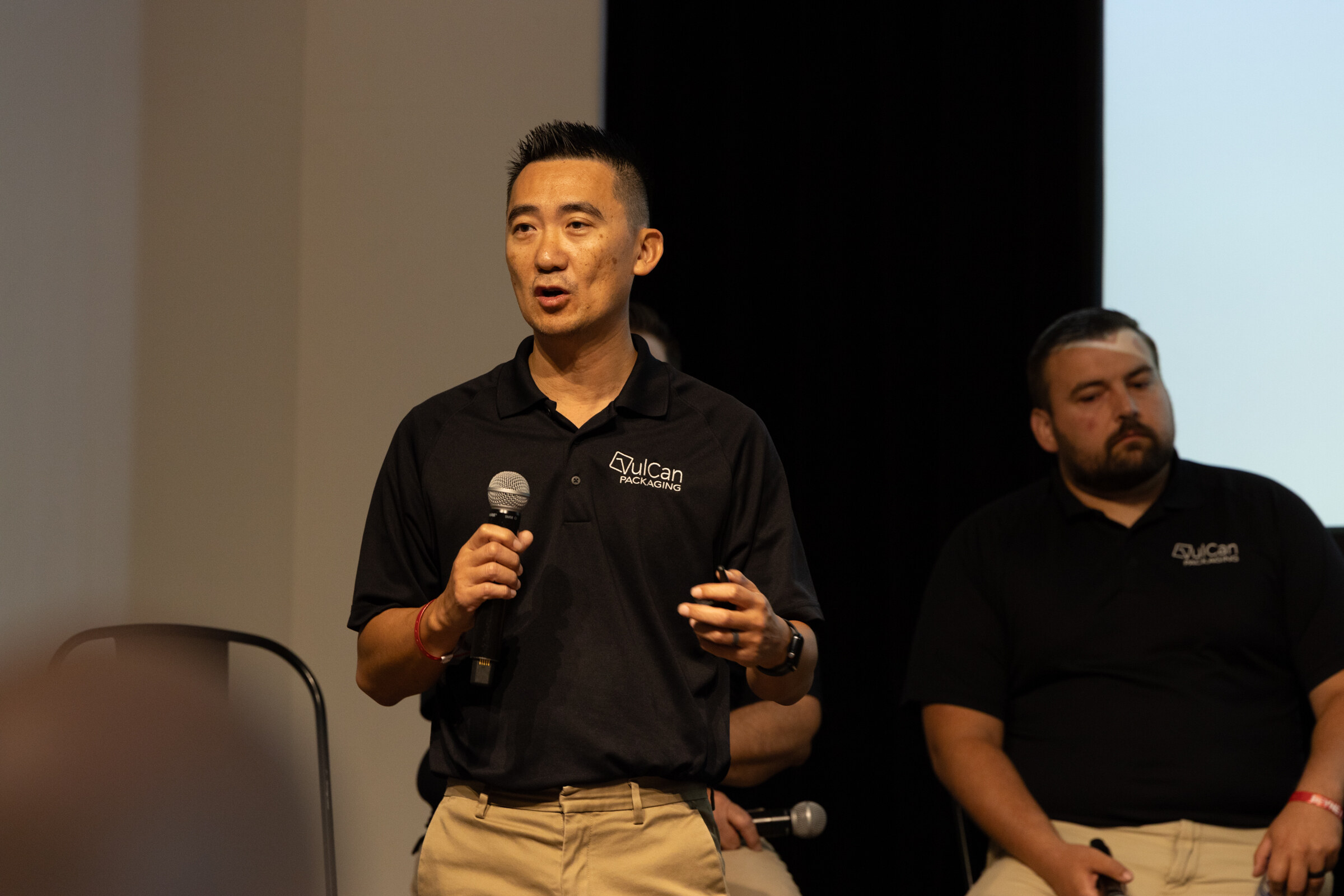 Man in a black VulCan Packaging polo speaking into a microphone, a colleague seated behind him.