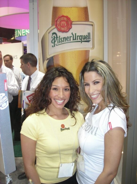Two smiling women pose in front of a large Pilsner Urquell beer glass graphic. One wears a yellow Pilsner Urquell shirt, the other a white Peroni shirt. Visitors stand in the background.