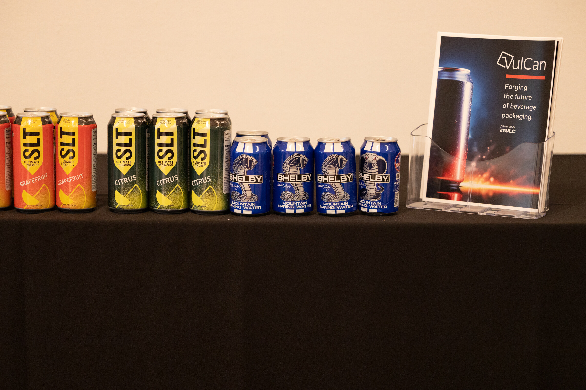 Row of SLT and Shelby beverage cans on a black table next to a VulCan brochure.