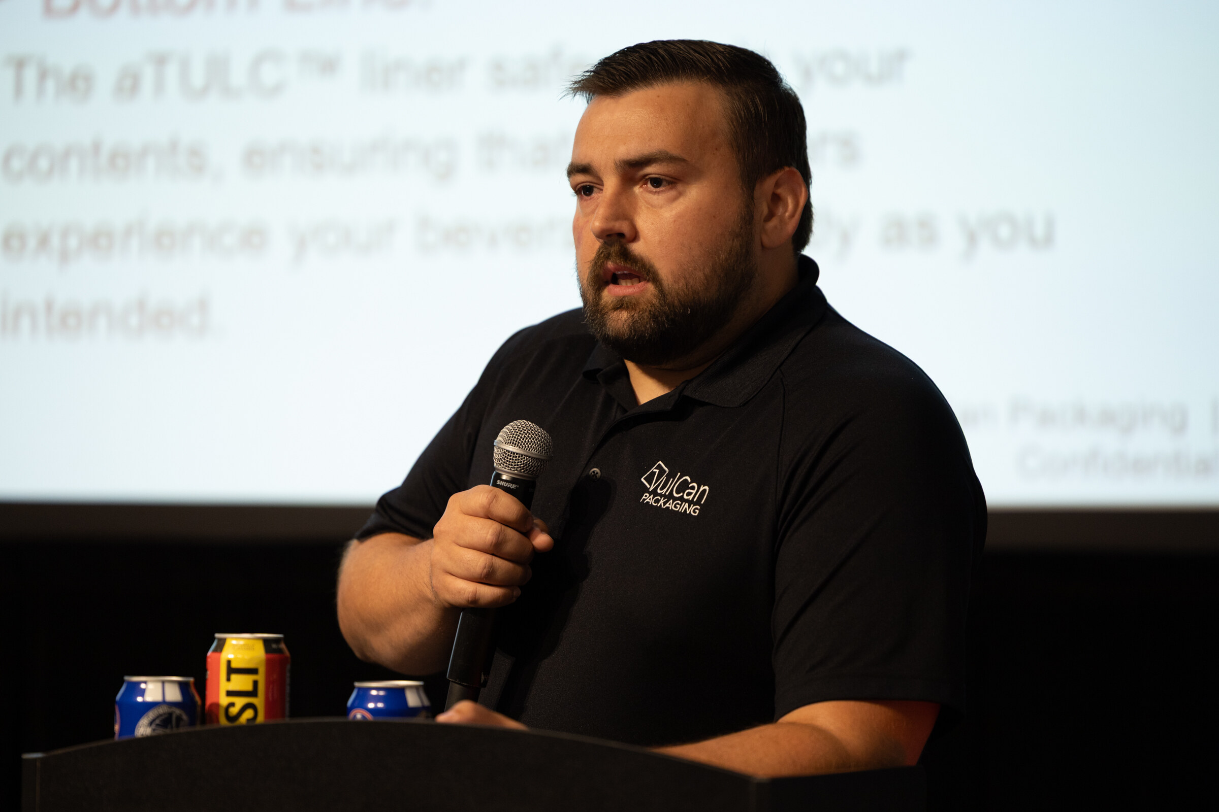 Man in a black VulCan Packaging polo speaking at a podium with cans in front of him and a slide about the aTULC liner behind.