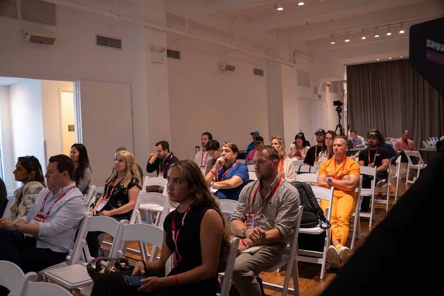 Seated attendees in rows of white folding chairs, one in an orange jumpsuit, facing the stage.