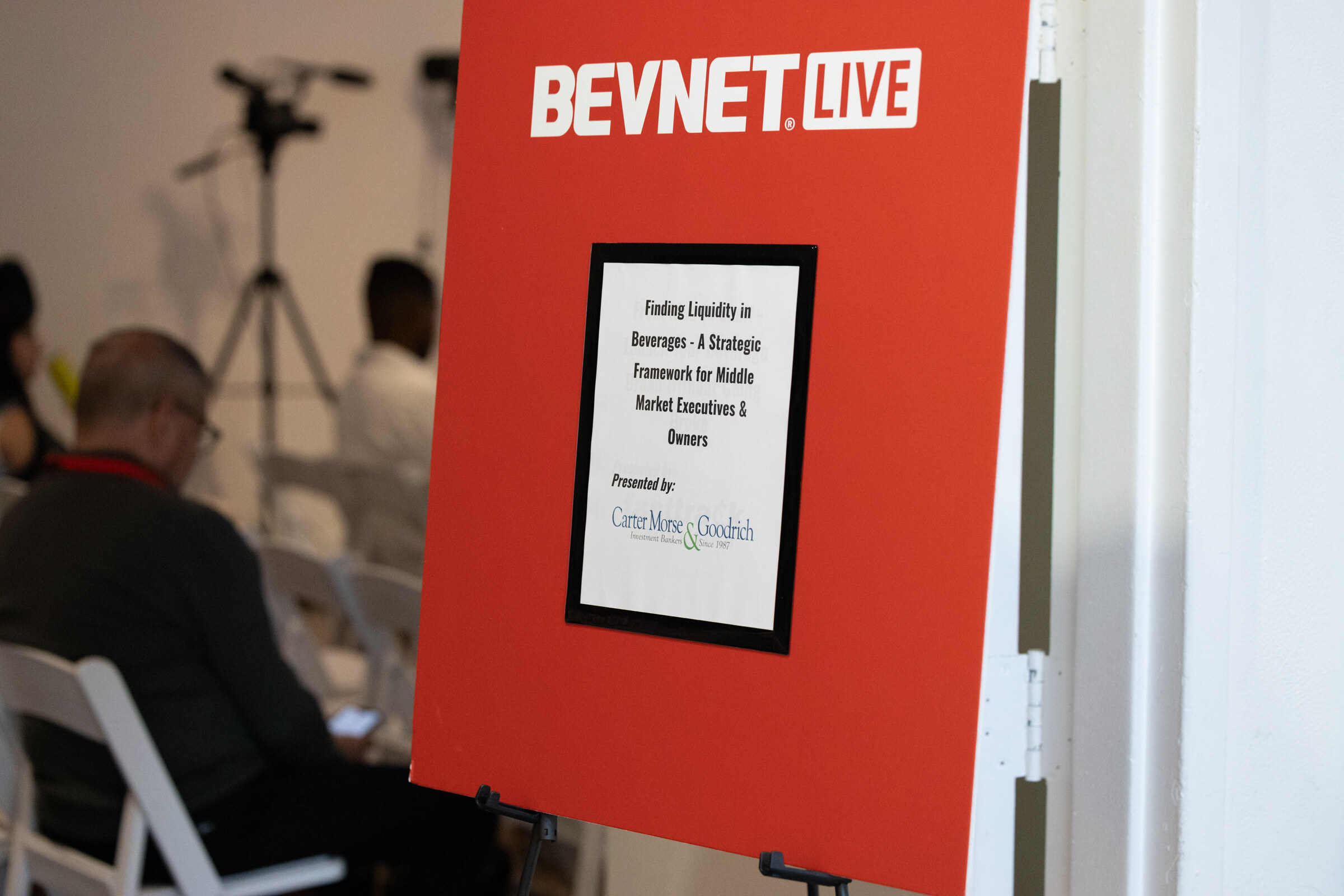 Red BevNET Live easel sign with the Carter Morse & Goodrich workshop title, attendees and a camera behind it.
