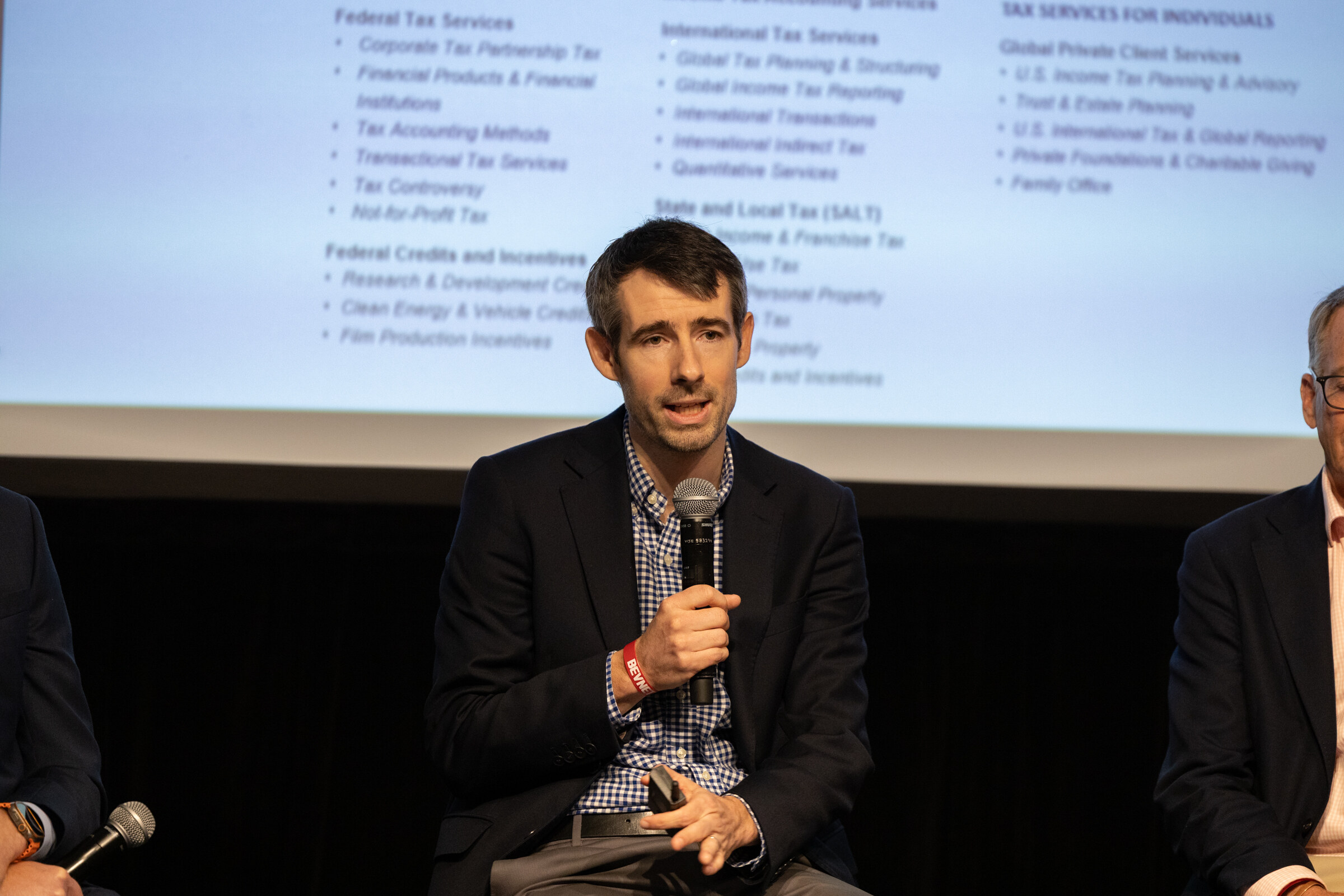 Man in a navy blazer and checked shirt speaking into a microphone in front of a services slide.