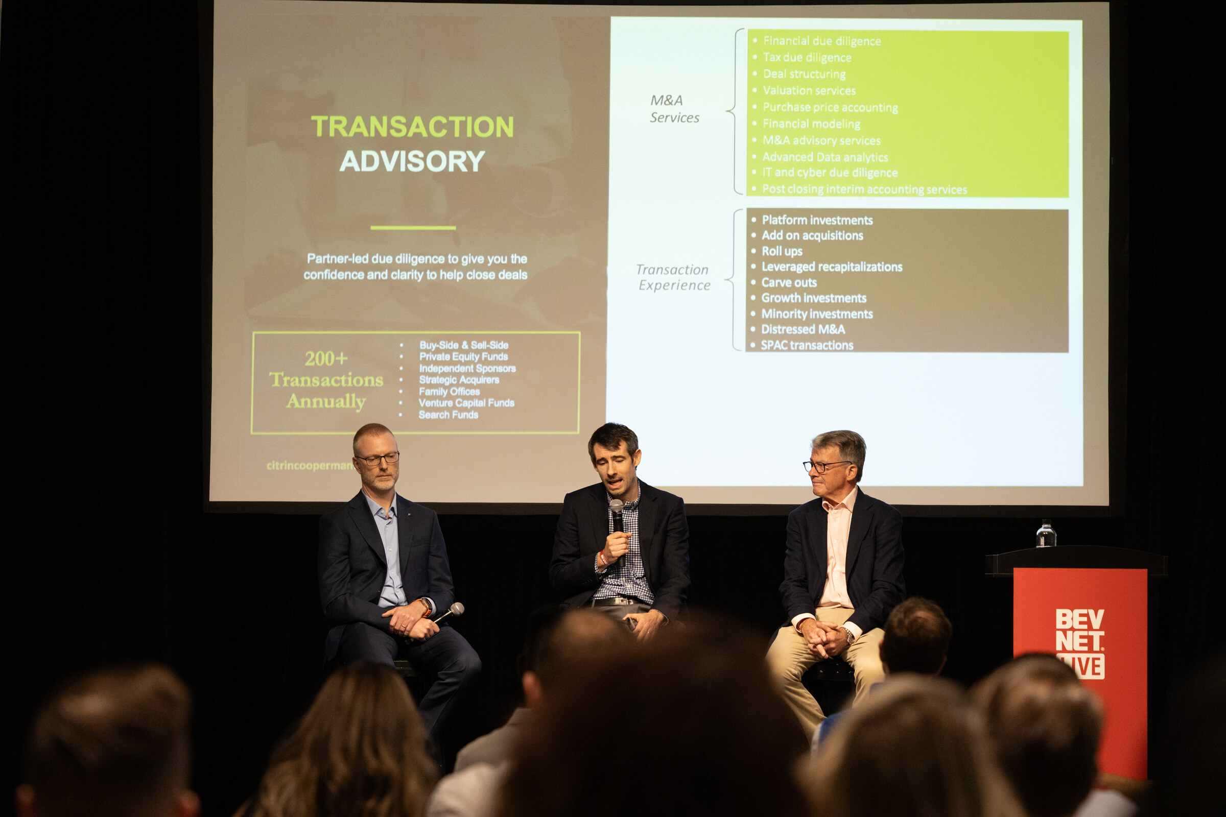Three panelists on stools beneath a “Transaction Advisory” slide, seen over the audience.