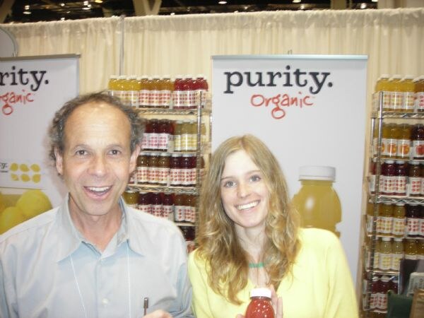 A man in a light blue shirt and a woman in a yellow top smile in front of Purity Organic banners. Shelves of juice bottles are behind them, and the woman holds a small bottle of red juice.
