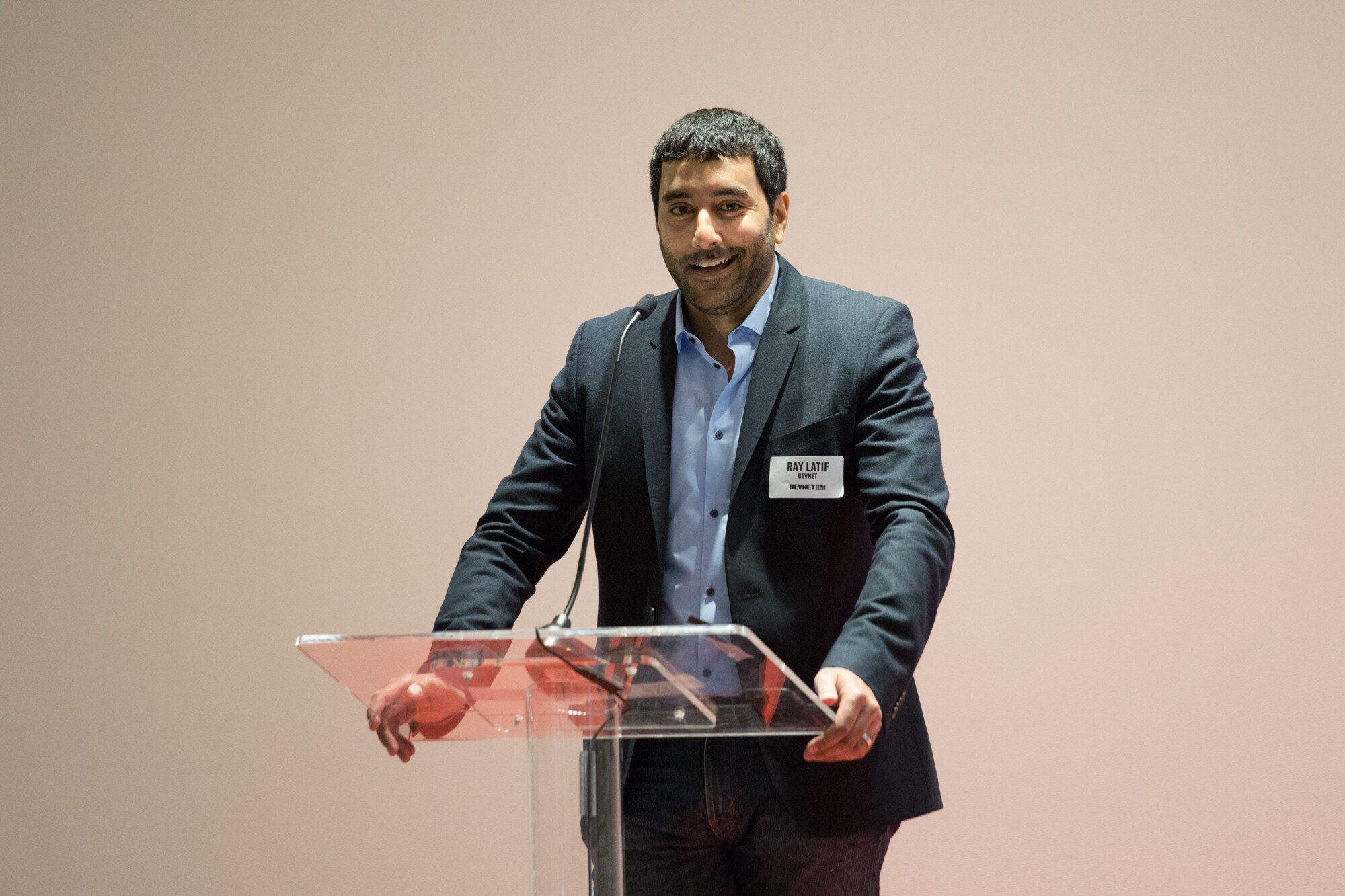 Man in a navy blazer and light blue shirt with a name badge smiling at a clear acrylic podium against a plain pink wall.