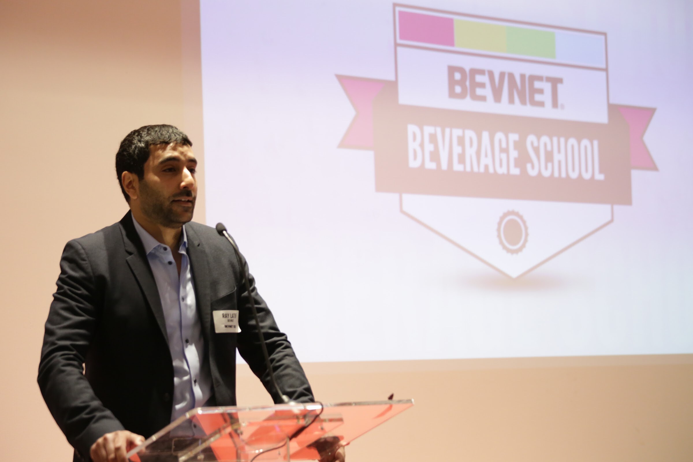 Man in a dark blazer and light blue shirt at a clear podium, a projection reading BevNET Beverage School with a pink ribbon and shield logo behind him.