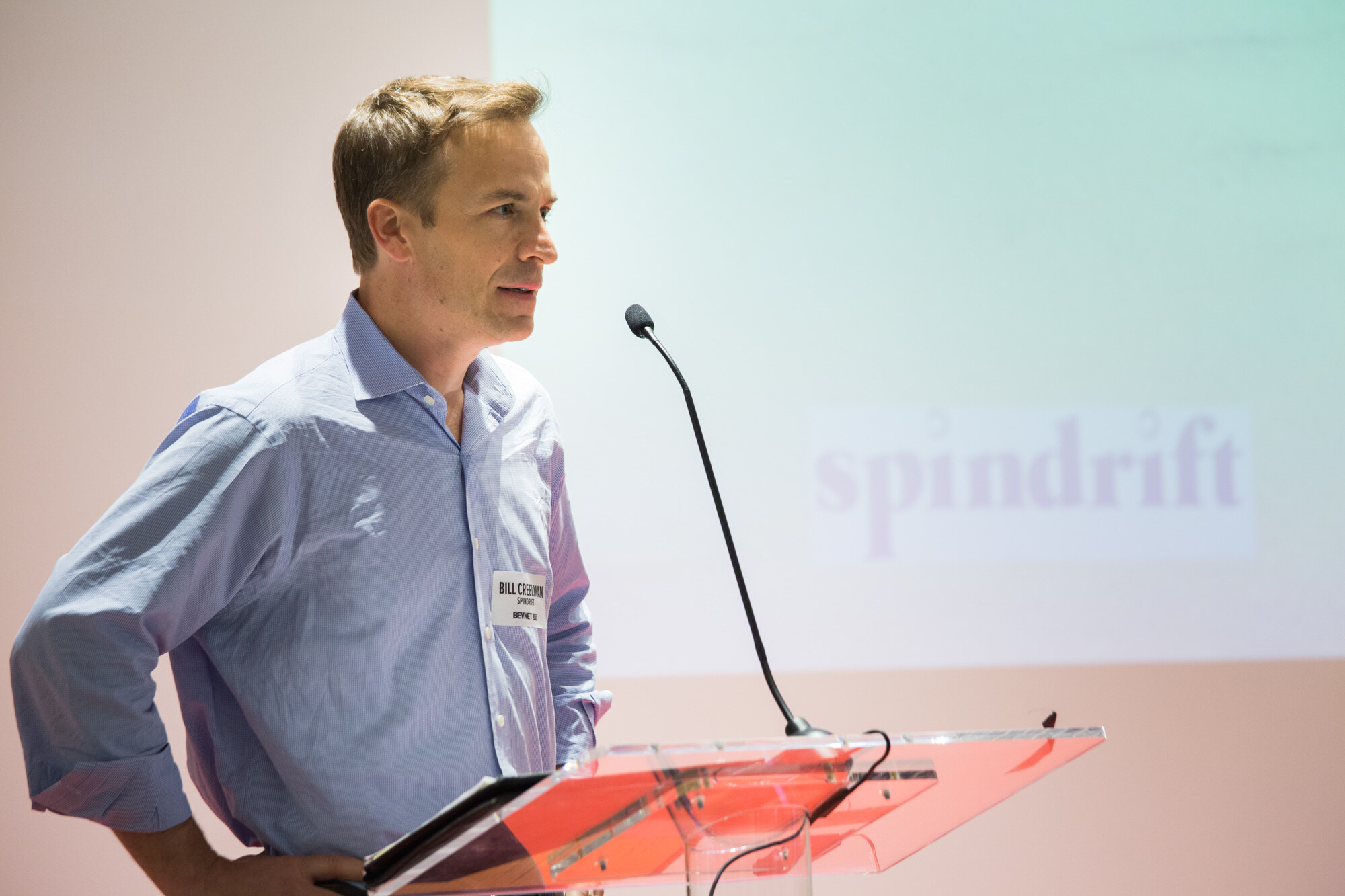 Man in a light blue button-up shirt with a name badge at a clear podium with a microphone, a projected slide with the word spindrift behind him.