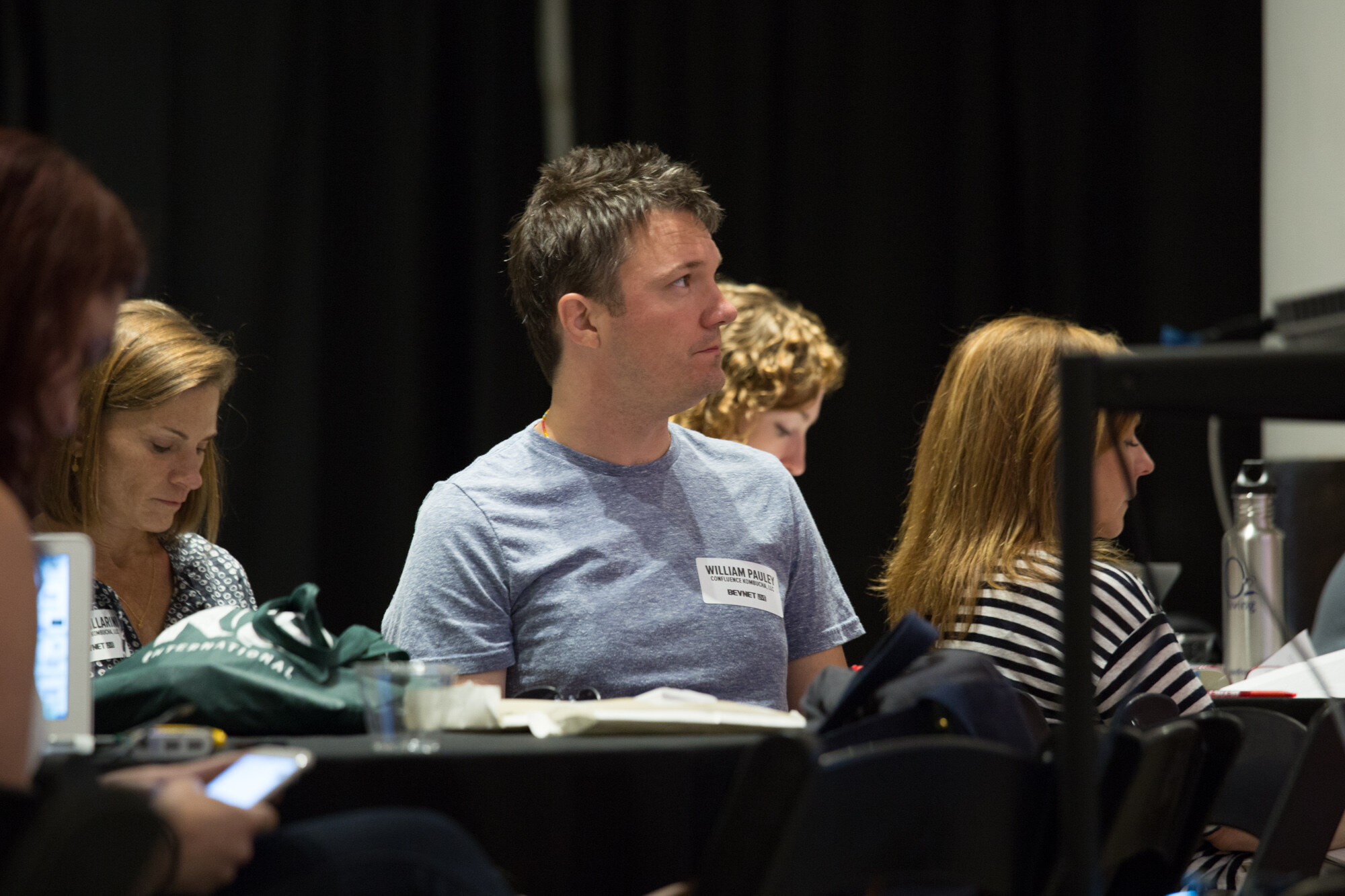Man in a grey t-shirt with a name badge seated at a black-draped table, with women taking notes beside him and a green tote bag on the table.