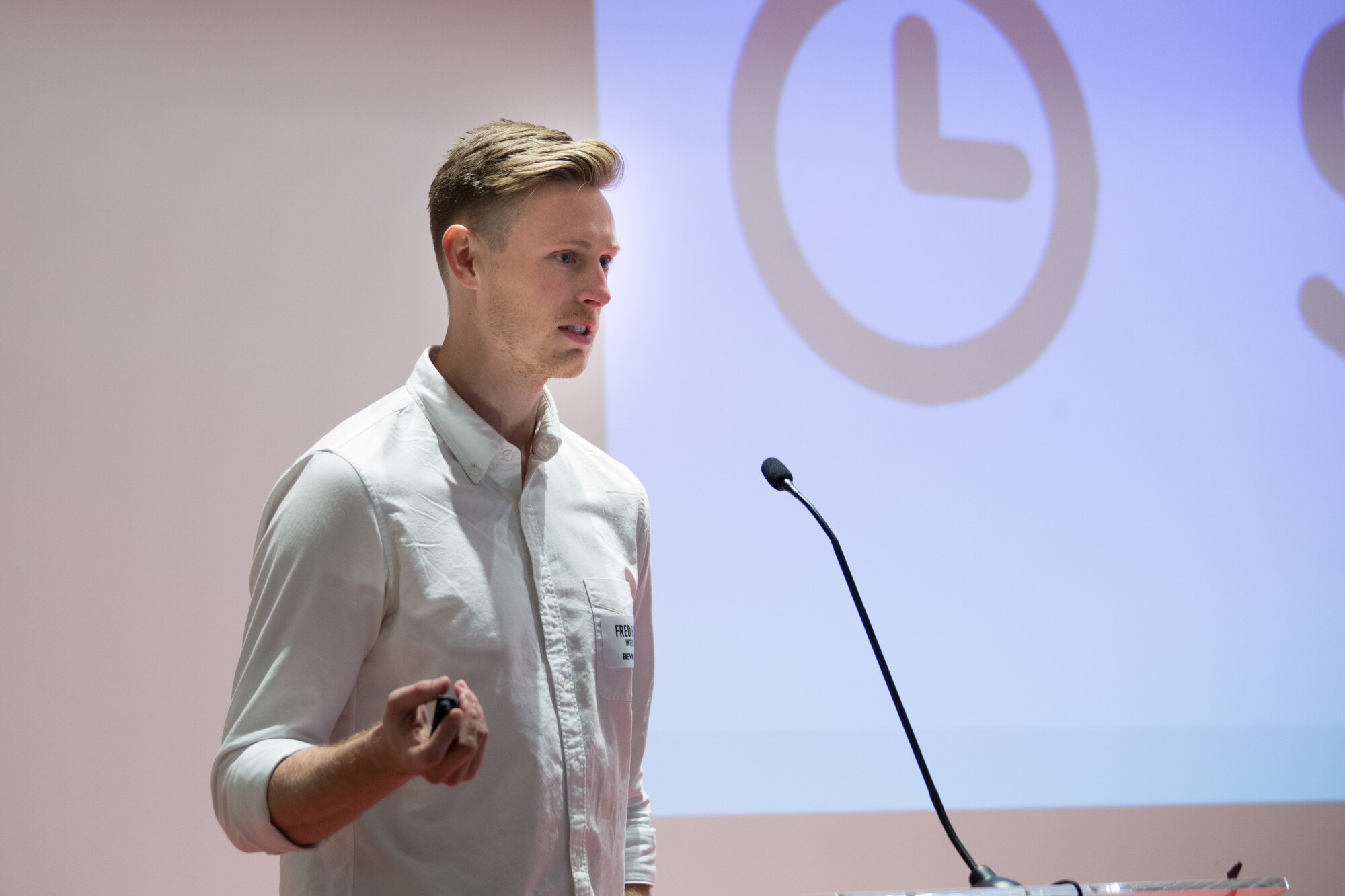 Man in a white button-up shirt with a name badge holding a presentation clicker beside a large projected clock icon.