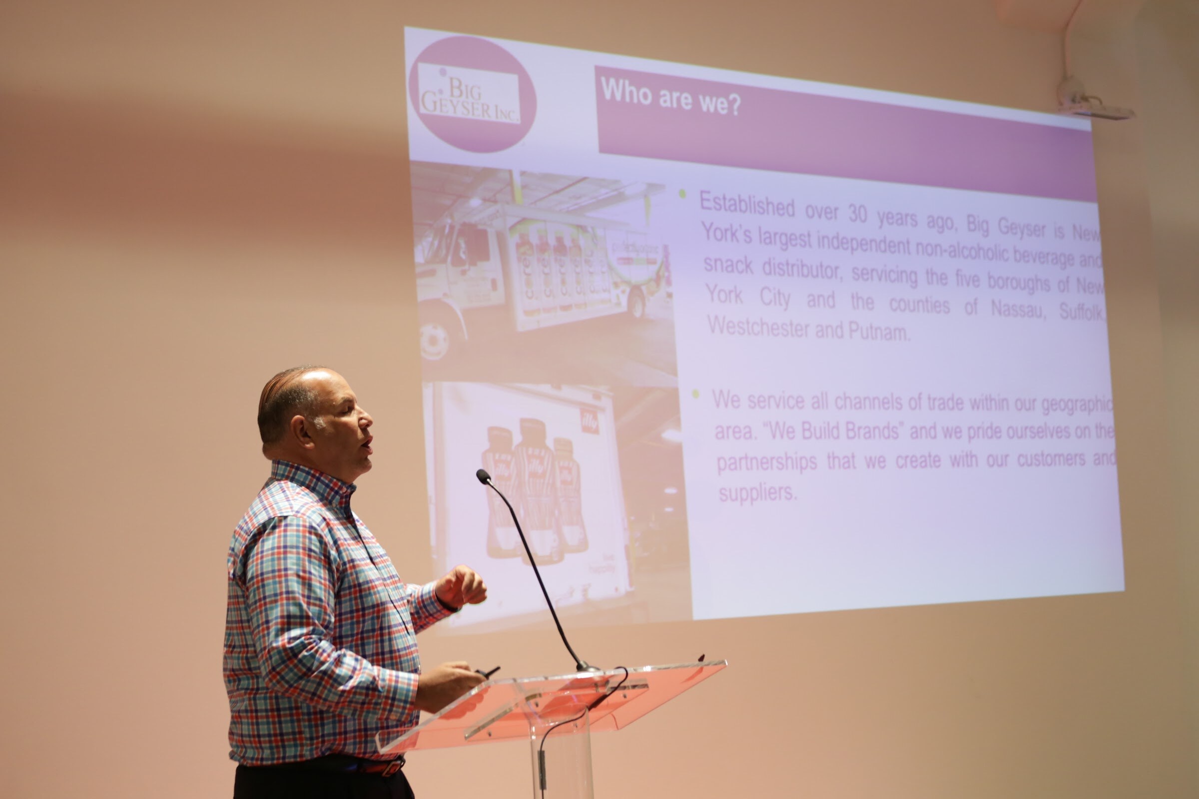 Man in a red, blue and white plaid shirt at a clear podium, a projected slide headed Who are we? with the Big Geyser Inc. logo and a photo of a delivery truck.