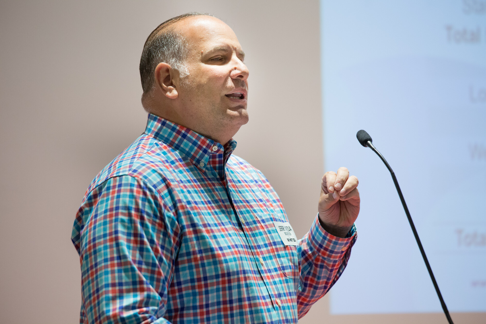 Close view of a man in a red, blue and teal plaid shirt with a name badge speaking beside a microphone, with a projected slide behind him.