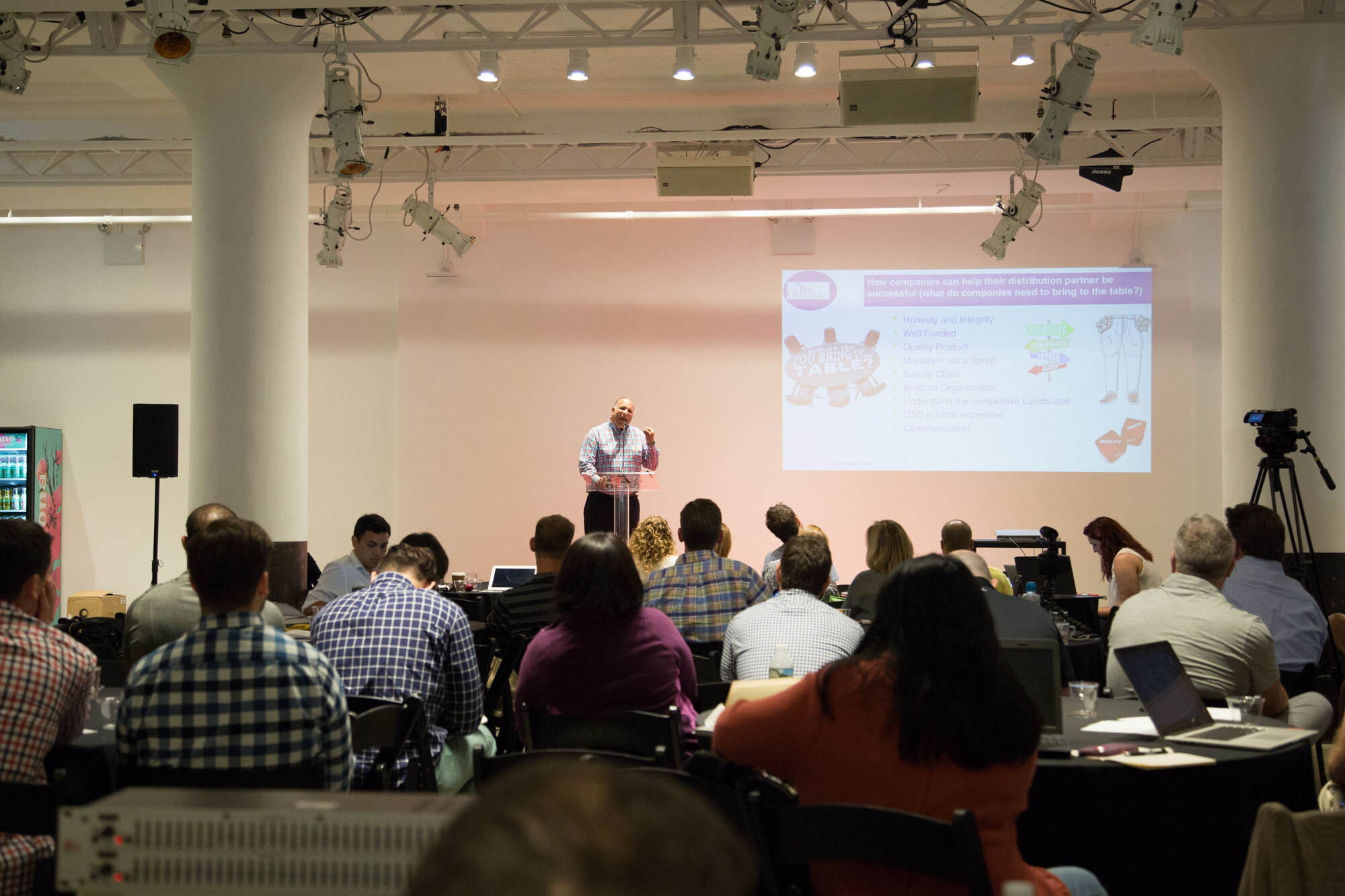 Wide view of the Beverage School room with a presenter at a podium, a screen headed How companies can help their distribution partner be successful, seated attendees with laptops and a video camera on a tripod.