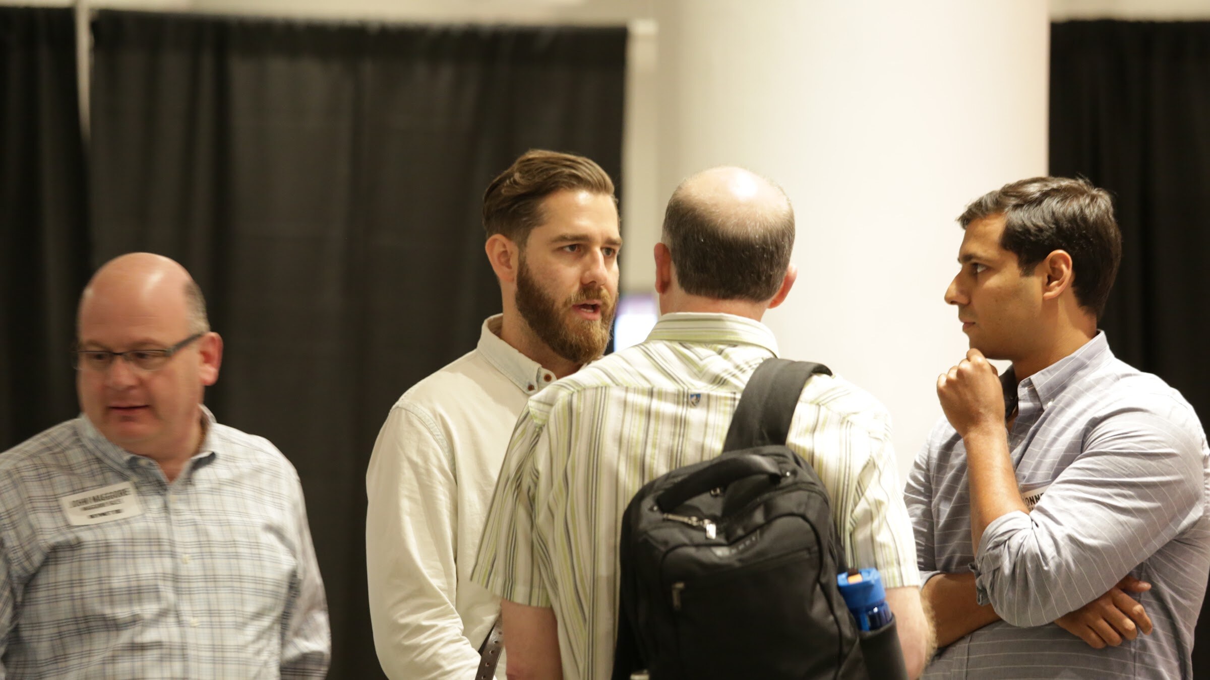 Four men in conversation in front of a white column and black curtain; a bearded man in a cream shirt speaks while another man with a black backpack faces away.