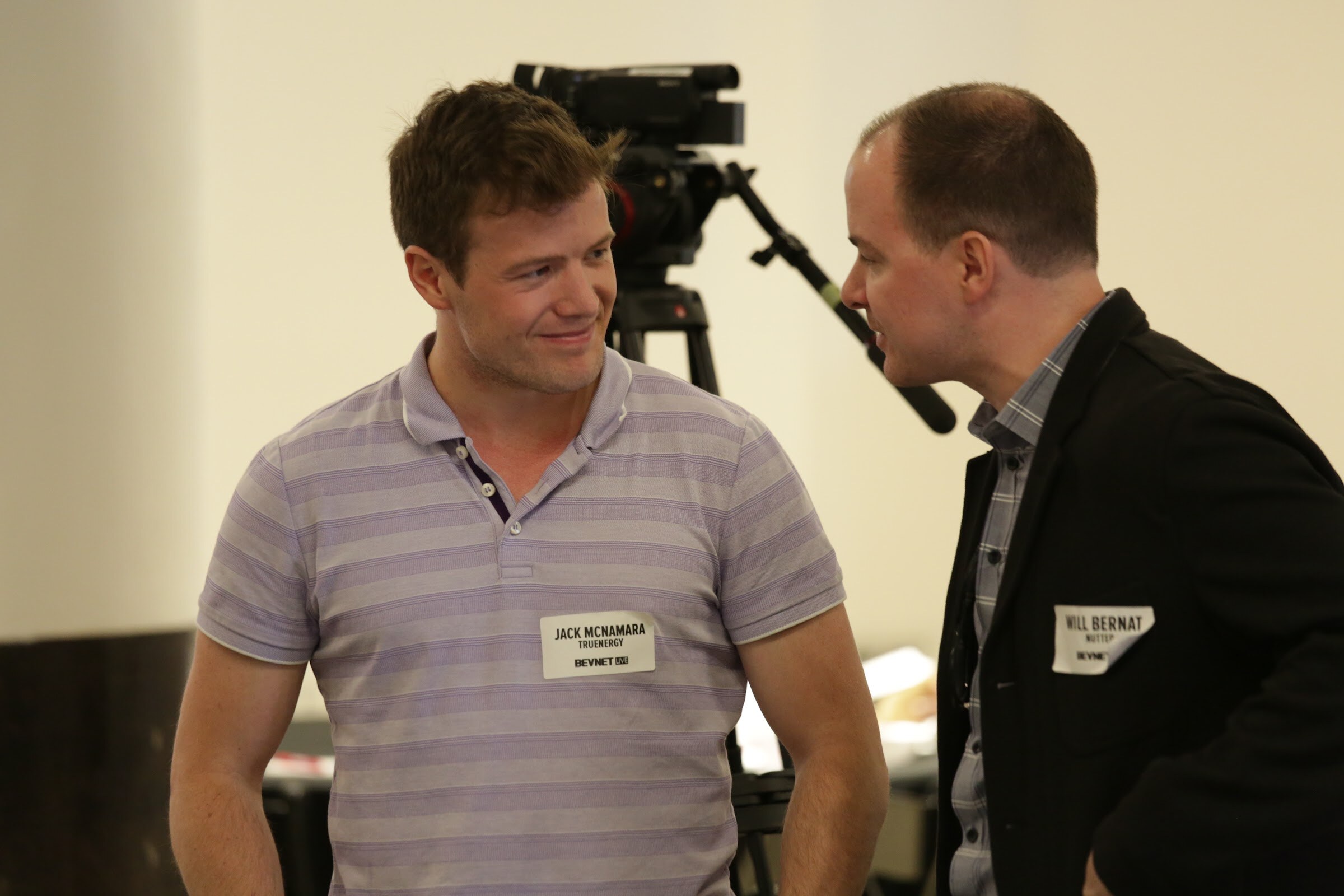 Man in a lavender striped polo smiling and looking sideways at a man in a black blazer speaking to him, with a camcorder on a tripod behind them.