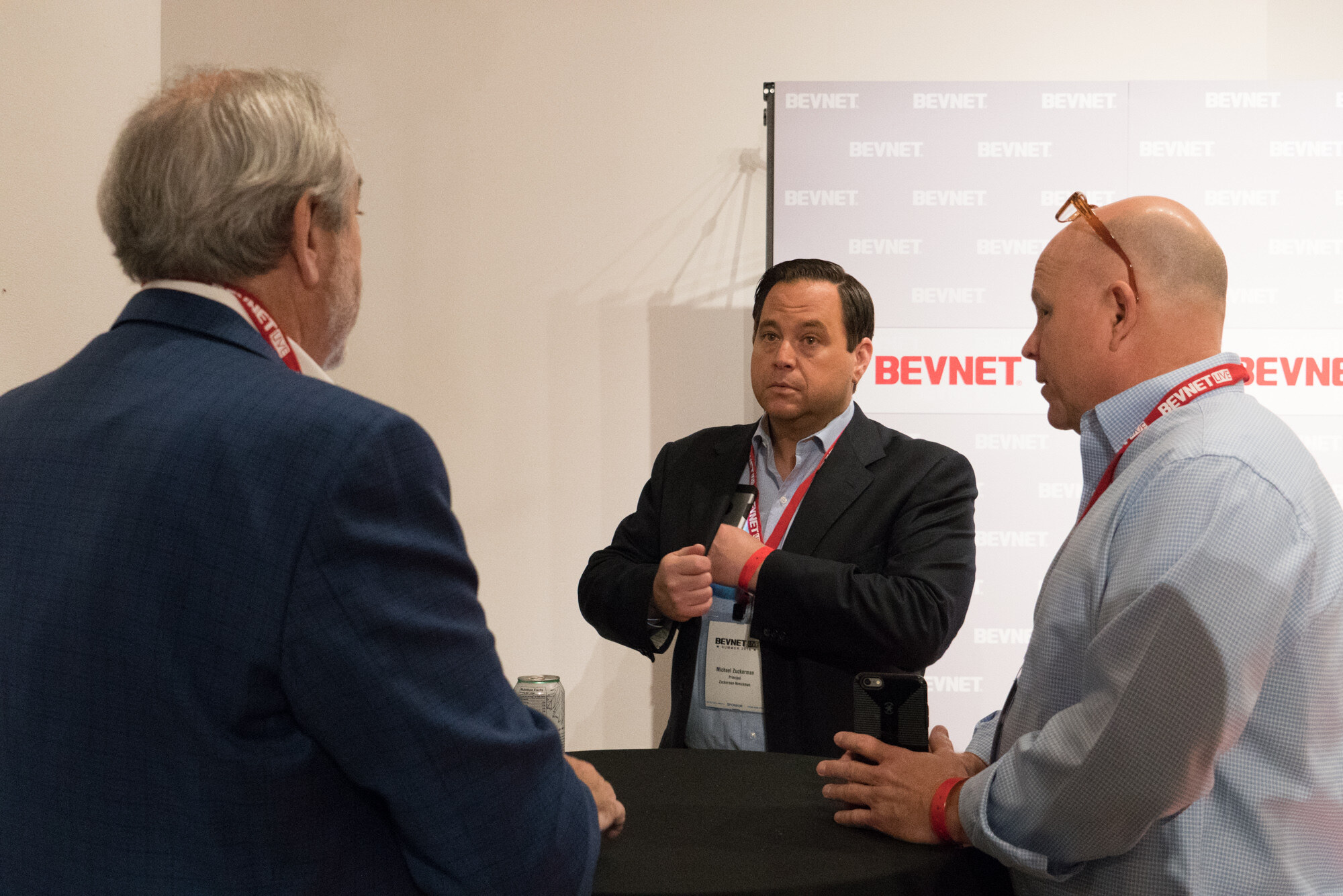 Three men with BevNET Live lanyards talking around a black high-top table in front of a BevNET step-and-repeat backdrop.