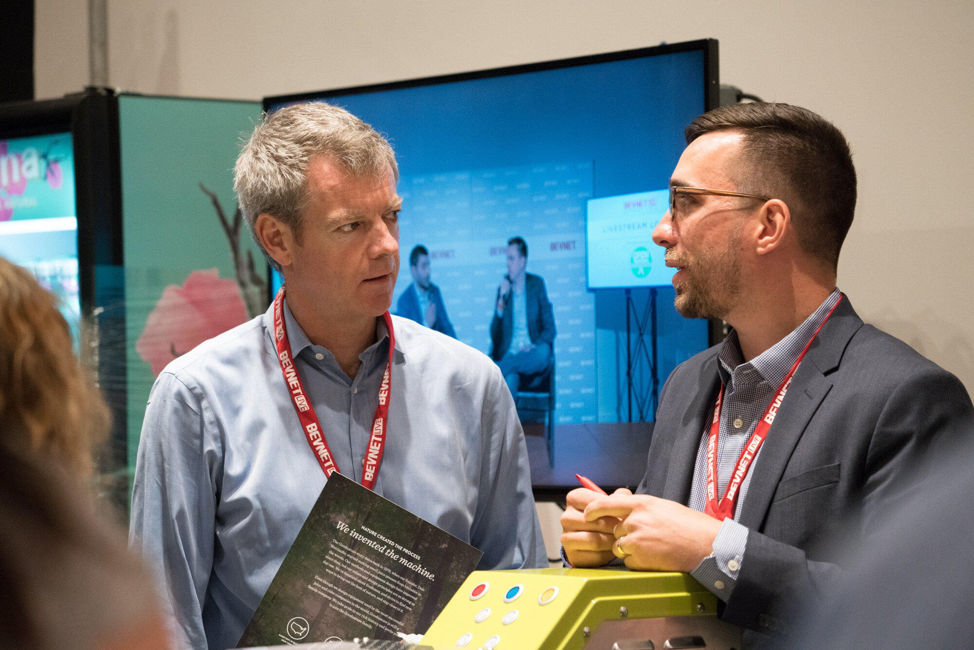 Two men with BevNET Live lanyards, one holding a brochure and the other a pen, in front of a monitor showing two people on the Livestream Lounge stage.