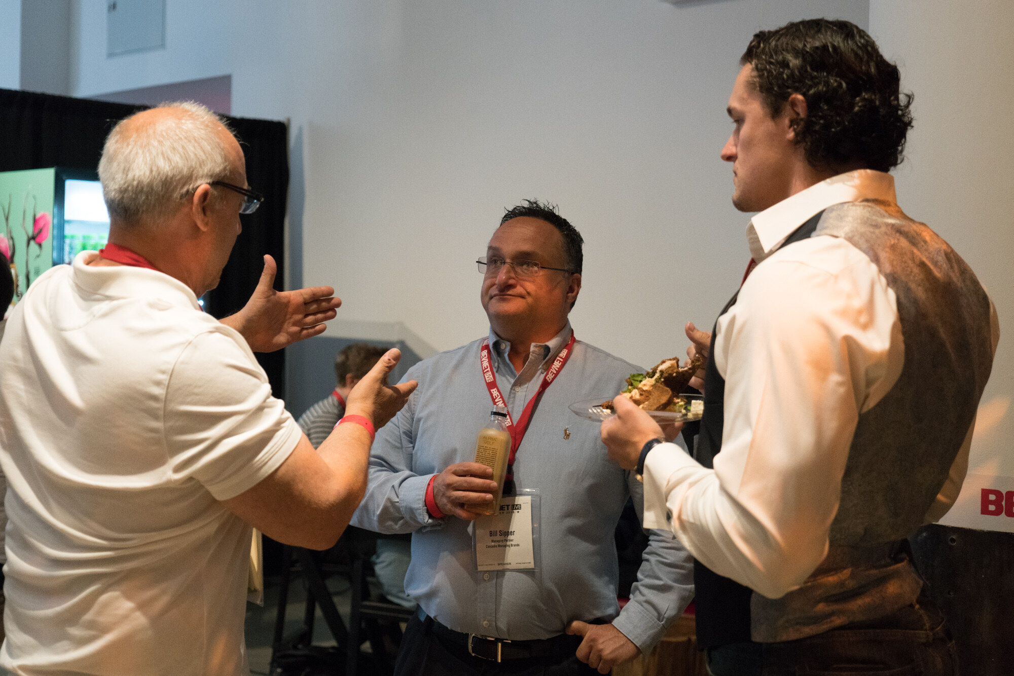A man in a white polo gesturing, a man in a light blue shirt holding a drink, and a man in a vest and white shirt holding a plate with a sandwich.