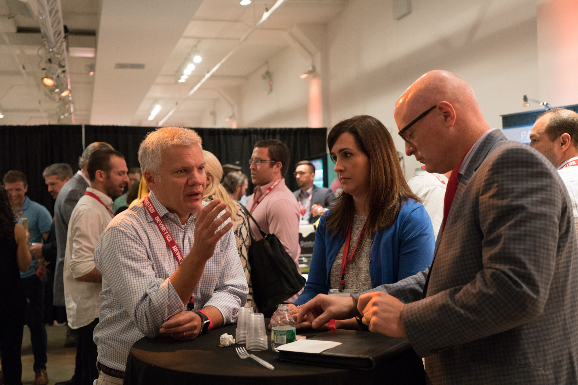 Three attendees with BevNET Live lanyards at a black high-top table with cups and a water bottle, with other attendees and black curtains behind them.