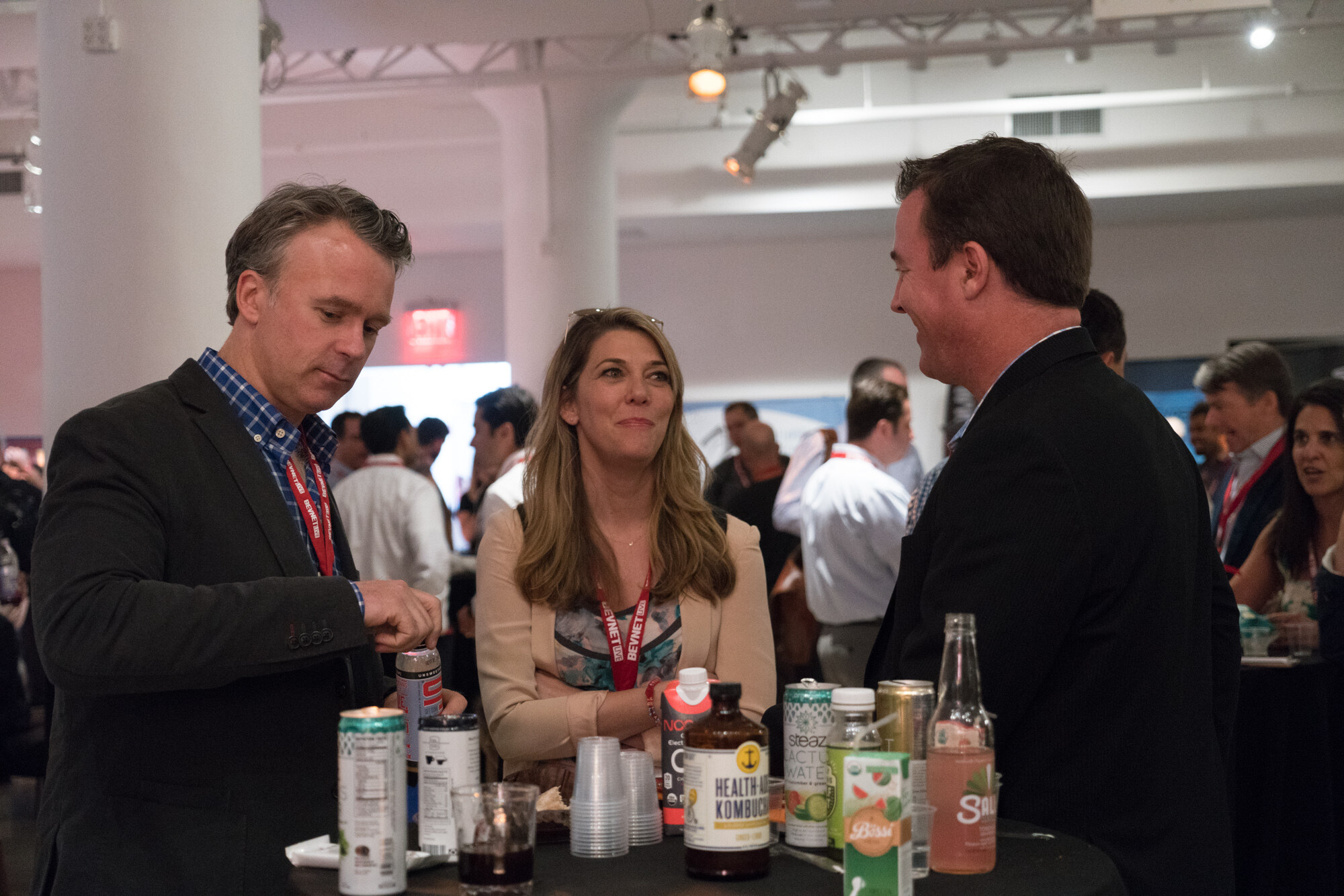 Three attendees in conversation around a black table with Health-Ade Kombucha, Steaz cans, a Bossa carton and cups, with other guests behind.