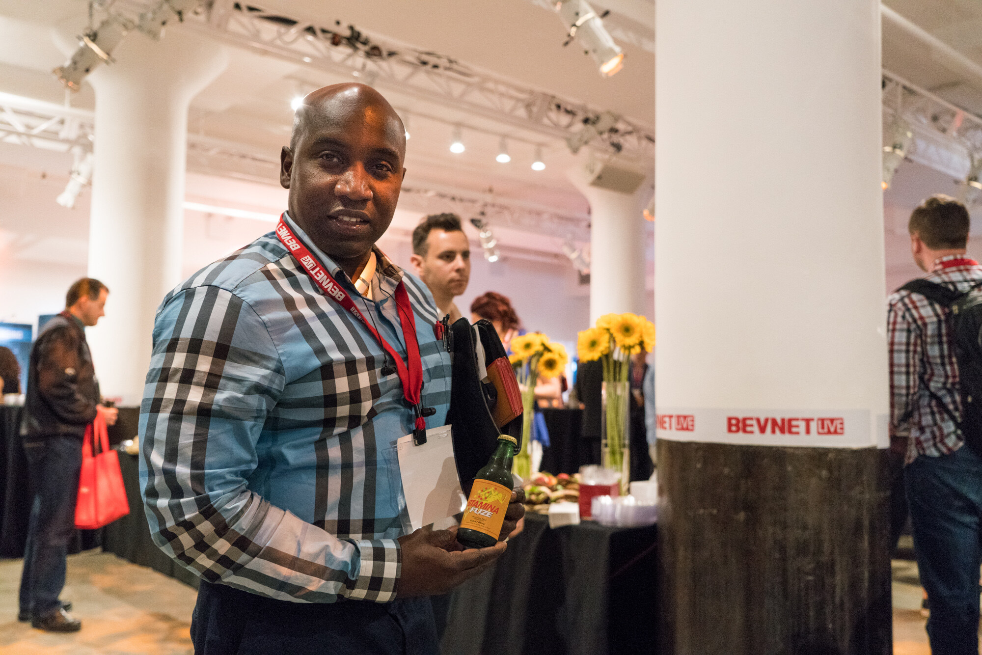 Man in a blue and black checked shirt with a BevNET Live lanyard holding a bottle labelled Vitamina, beside a white column with a BevNET Live band, sunflowers and a buffet table behind him.