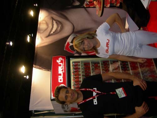 Sideways photo of a man in a black Guru shirt with a badge and a woman in a white Guru shirt, standing at a booth with a red Guru sign and a cooler of drinks.