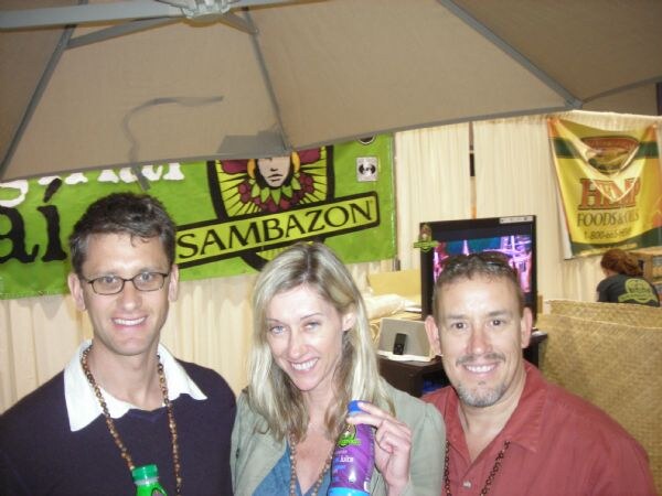 Two men and a woman smile in front of a green Sambazon banner. The woman holds a purple bottle, and a man at left holds a green-capped bottle near the bottom of the frame.