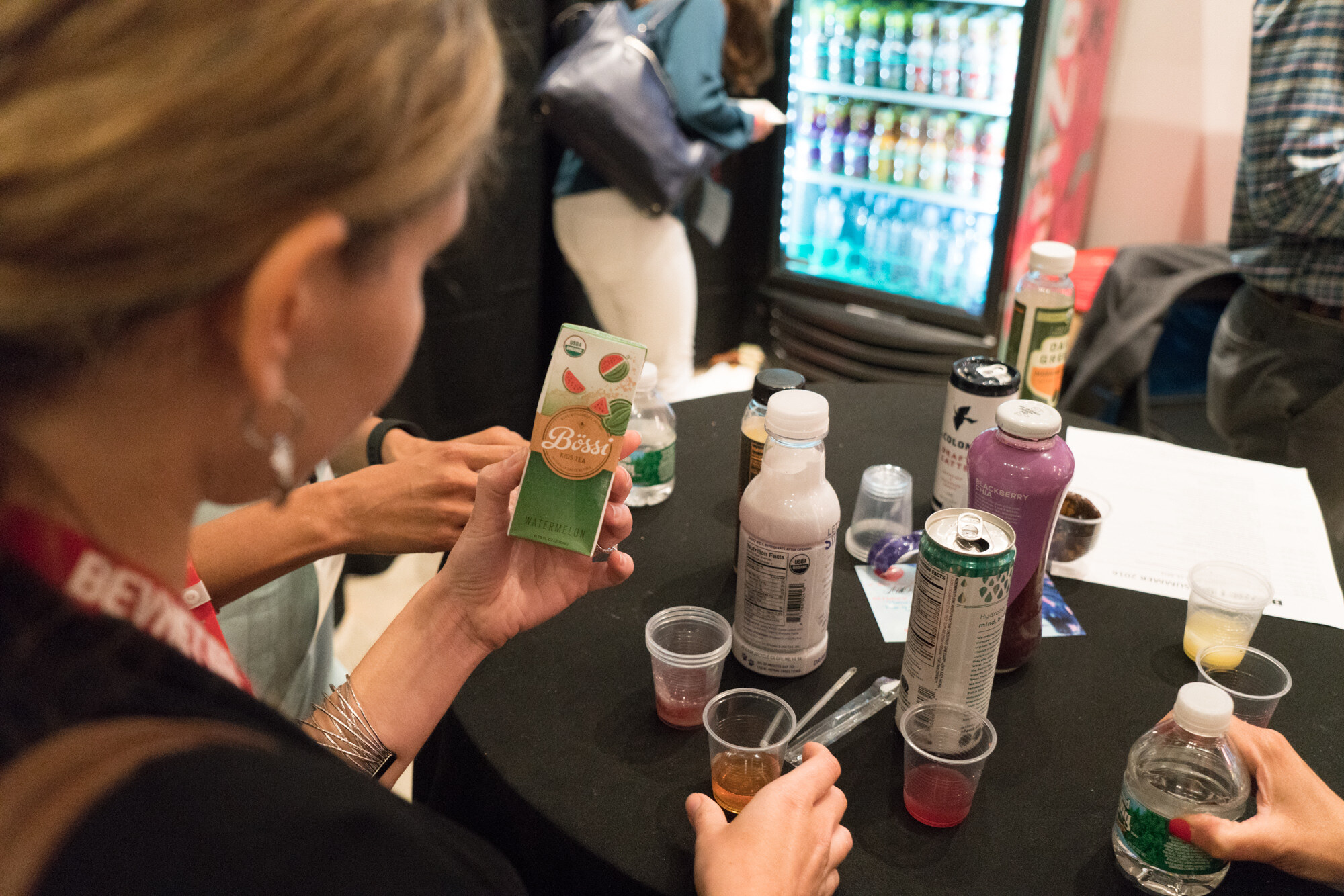 Close view of a woman in a BevNET Live lanyard holding a watermelon Bossa carton, with sample cups, a blackberry drink, a Daily Greens bottle and a can on a black table.