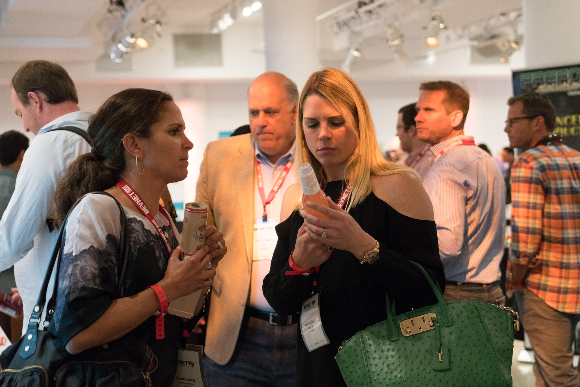 Women with lanyards examining a bottle and a can, a man in a tan blazer behind them and others in conversation in a crowded room.
