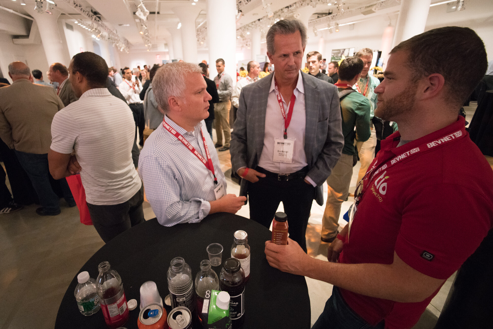 Wide-angle view of three men around a black table with bottled drinks and cups, one in a red Tio polo, with a crowded room behind them.