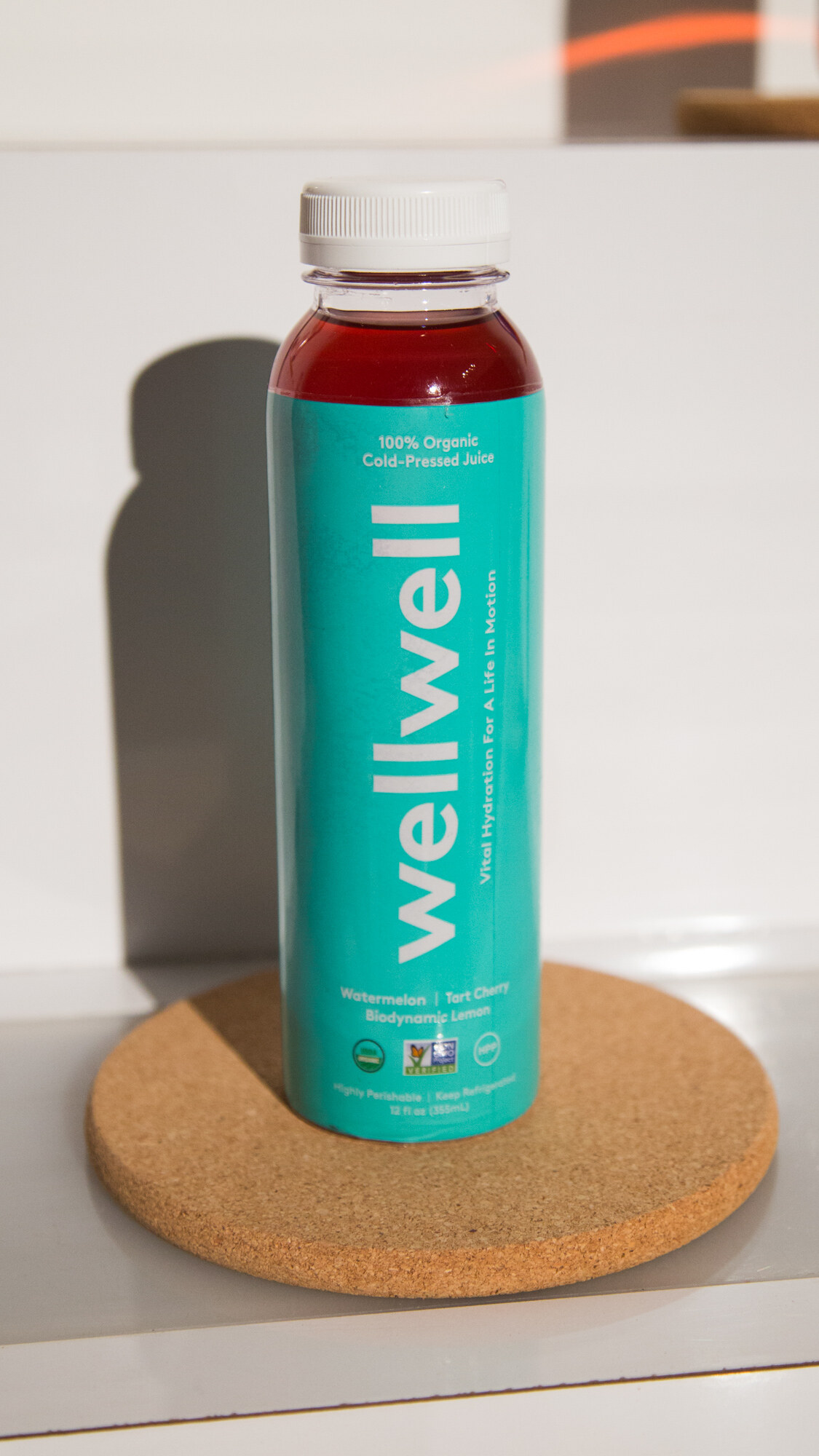 Teal-labelled wellwell bottle of 100 percent organic cold-pressed juice, watermelon and tart cherry, on a round cork coaster in front of a white shelf.