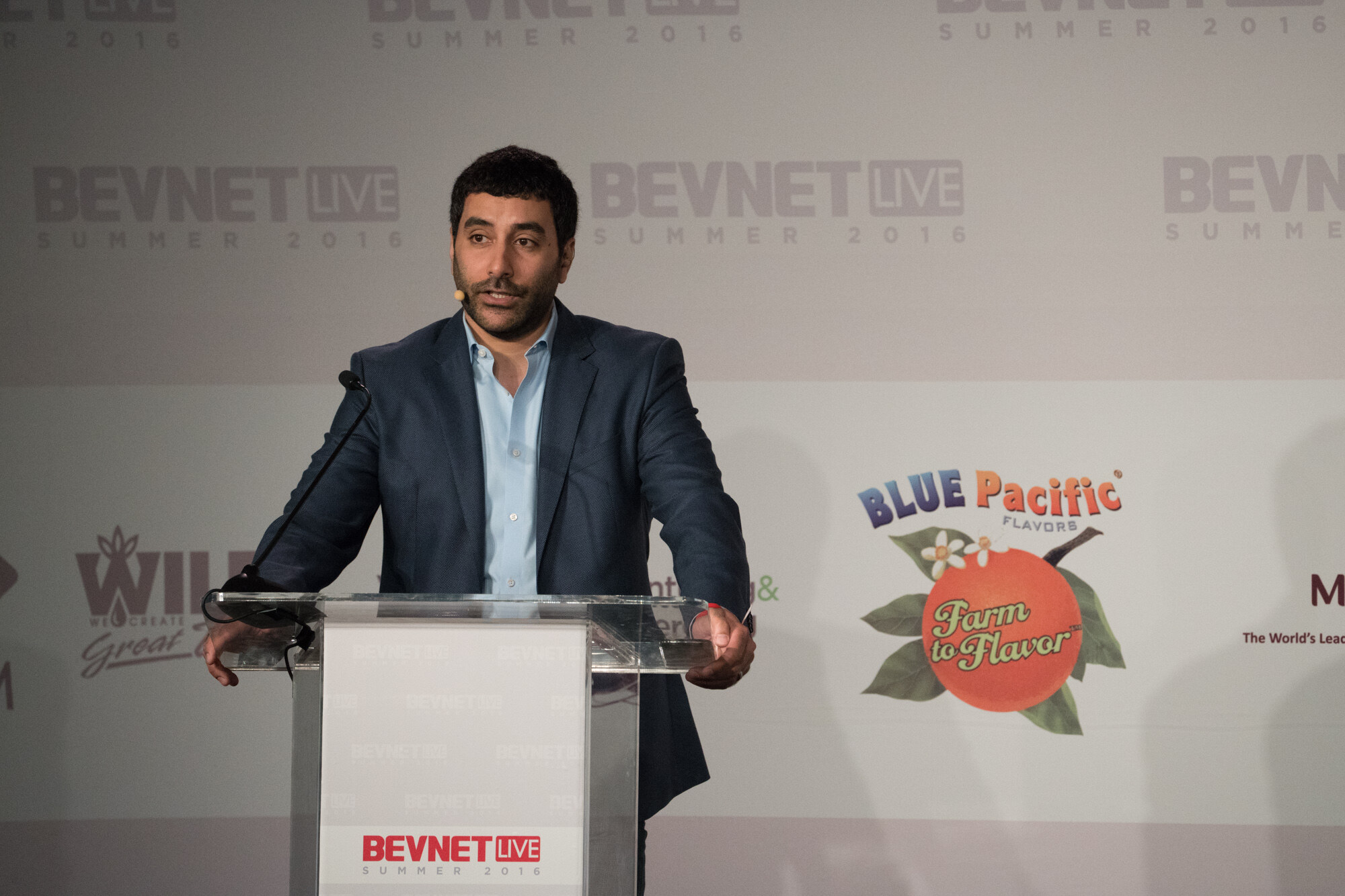 Man in a navy blazer and light blue shirt with a headset mic at a clear podium reading BevNET Live Summer 2016, with Blue Pacific Flavors and Wild logos on the backdrop.