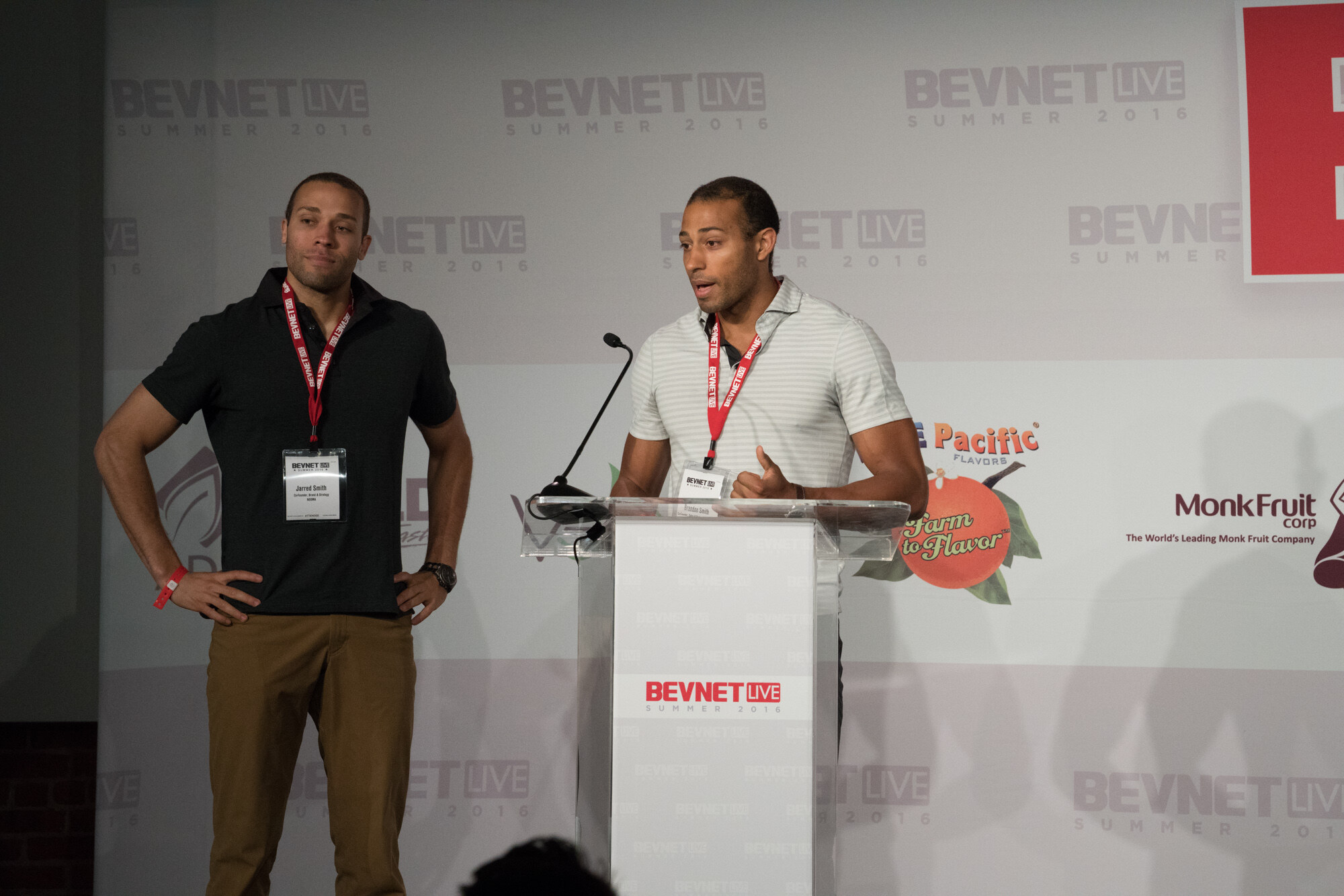 Two men in BevNET Live lanyards, one in a black polo and one in a striped polo, at a clear podium reading BevNET Live Summer 2016, with a Blue Pacific Flavors logo behind them.